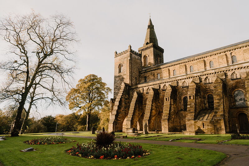 A large sandstone abbey with a square battlemented tower rises behind a circular flower bed in full bloom, an autumn tree standing beside the lawn in front.