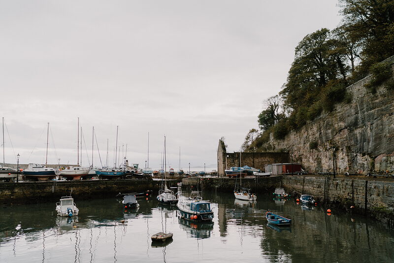 Yachts and small boats lie moored in a sheltered harbour beneath a sandstone cliff, a ruined stone wall running along the quayside.