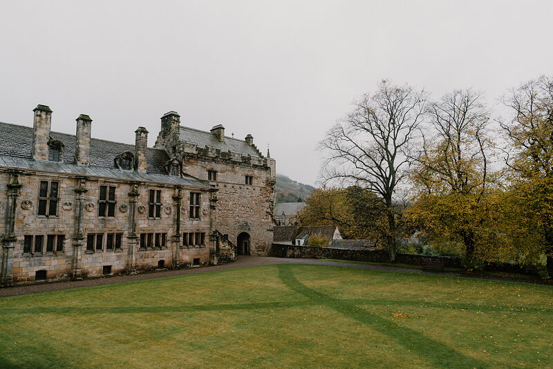 A turreted stone palace with tall chimneys overlooks a mown lawn, autumn trees glowing gold against the grey stonework.