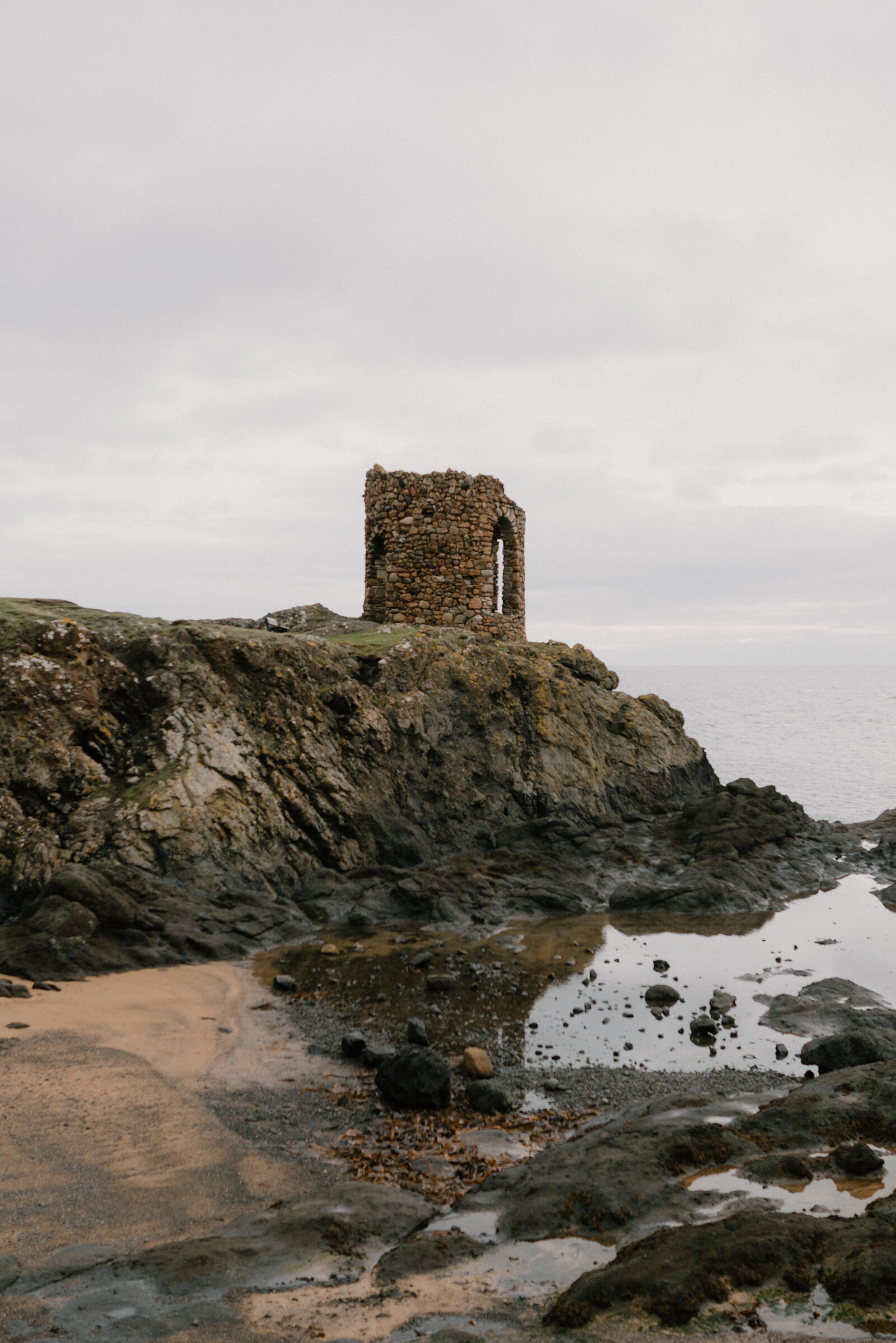 A ruined stone tower with an arched doorway stands on a rocky headland, tide pools reflecting its shape below.