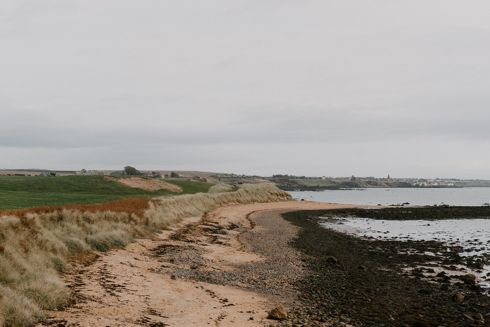 A sandy beach path curves along wind-blown dune grass, a distant town and harbour visible along the coastline.
