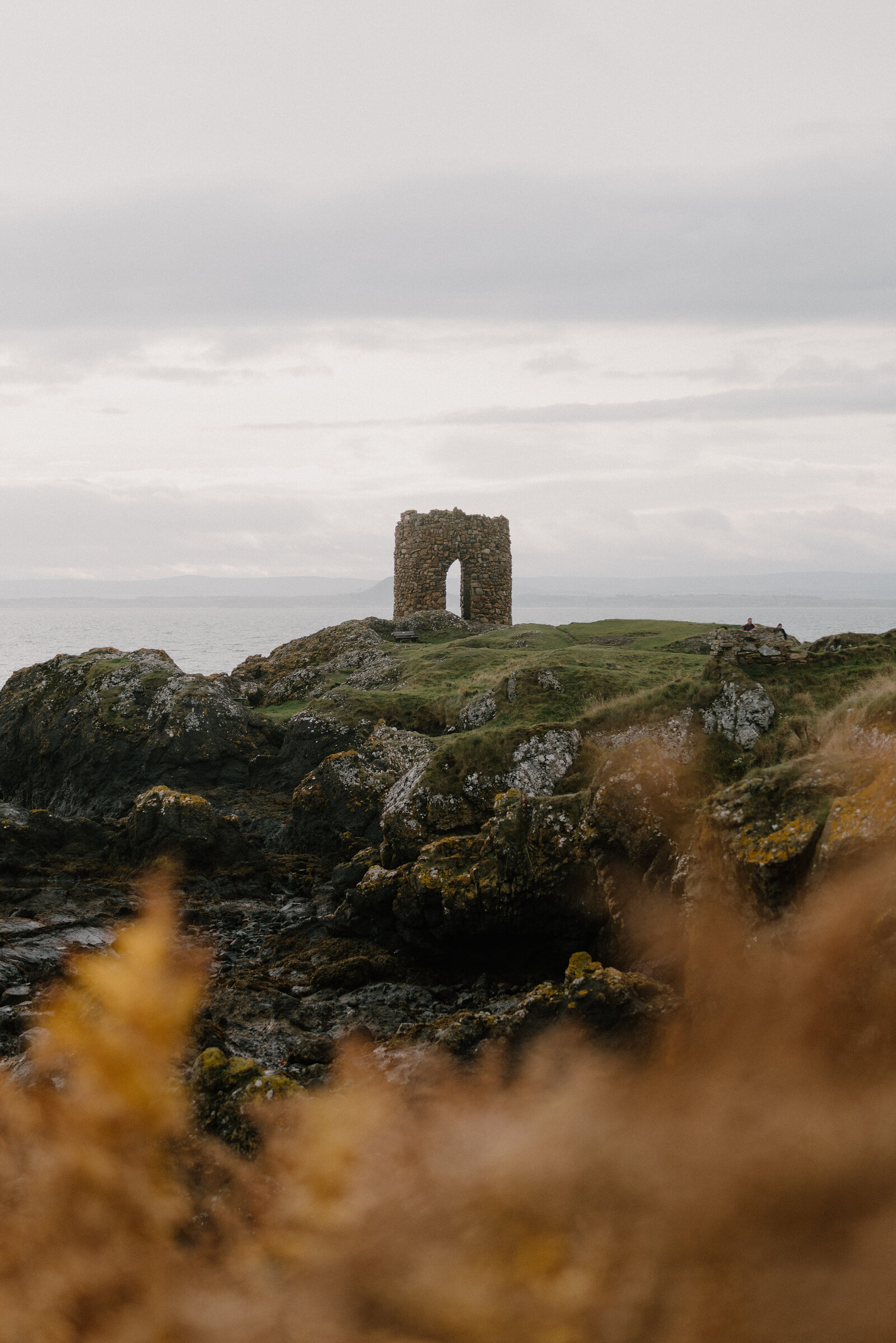 A ruined stone tower with an arched doorway stands on a rocky headland, autumn bracken framing the view in the foreground.