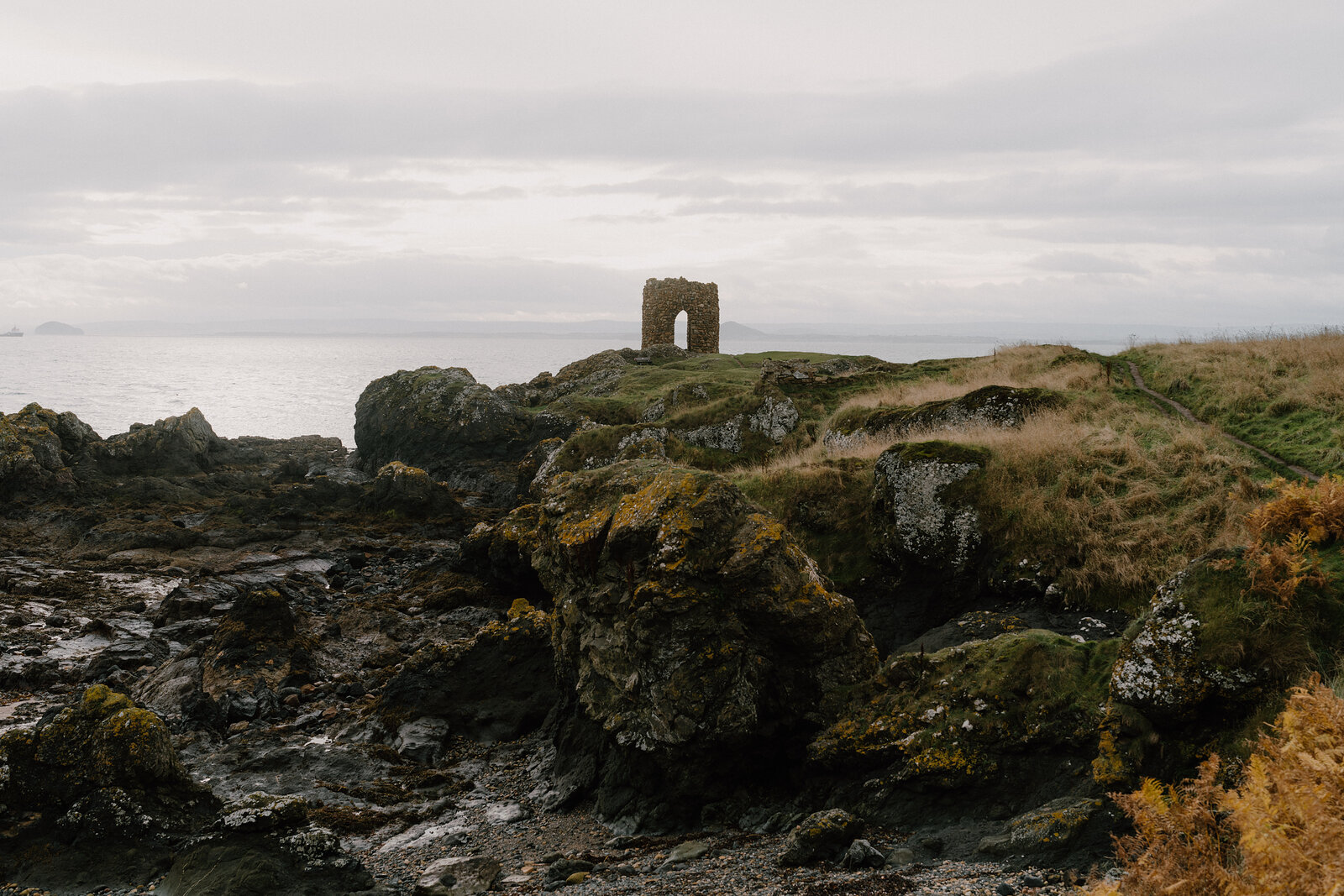 A ruined stone tower with an arched doorway stands on a mossy, rocky headland above the sea.