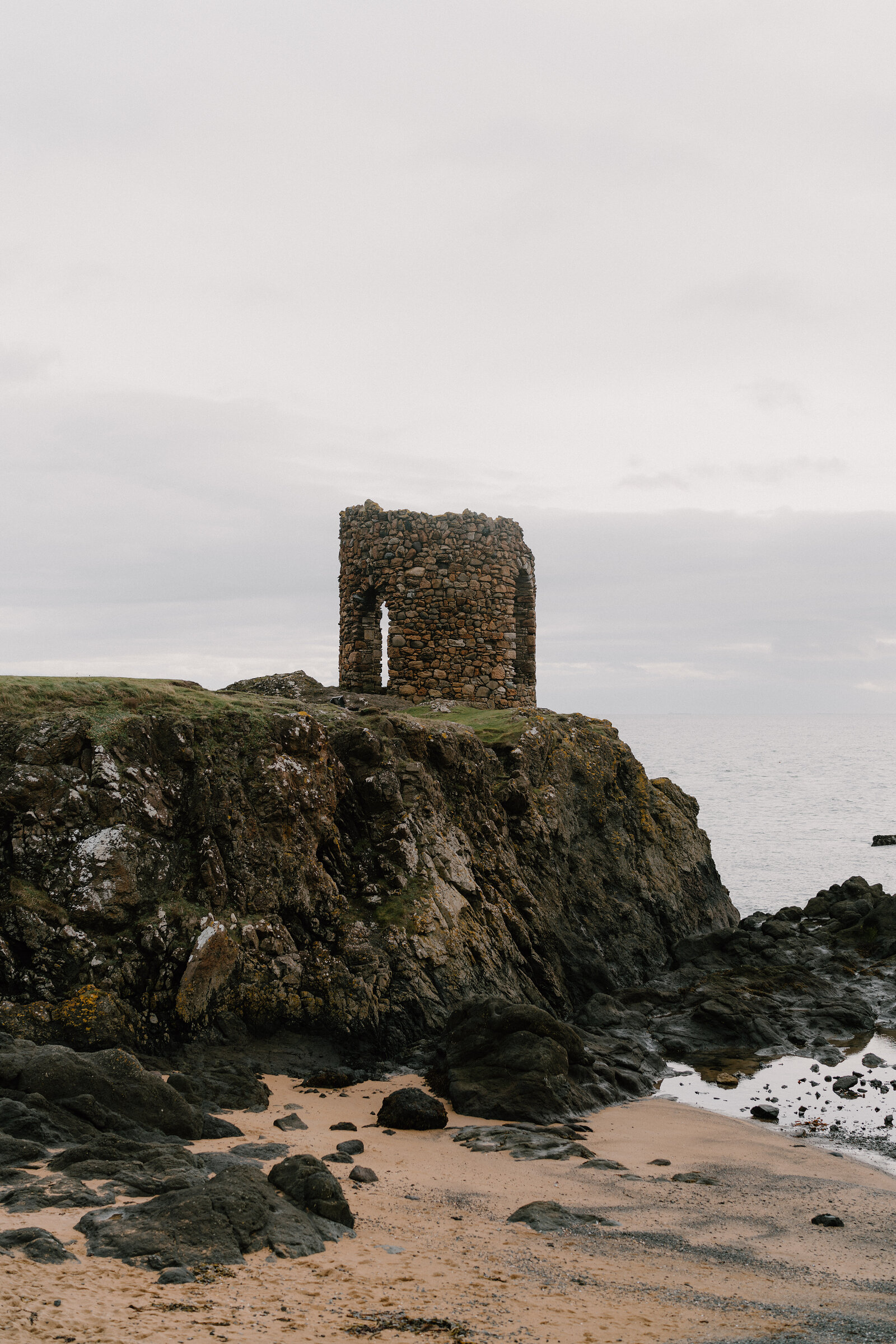 A ruined stone tower with an arched doorway stands on a rocky headland above a sandy beach, calm sea stretching behind.