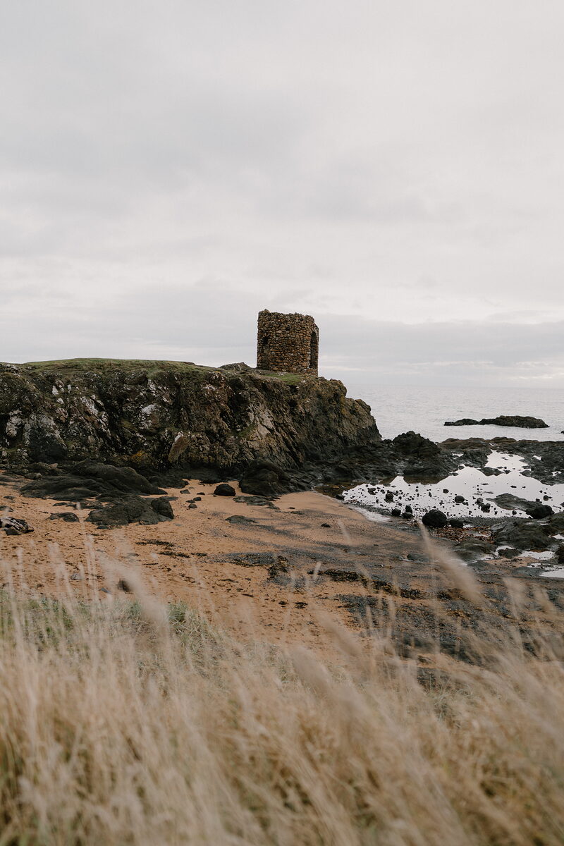 A ruined stone tower with an arched doorway stands on a rocky headland above a sandy beach, autumn grass framing the view in the foreground.