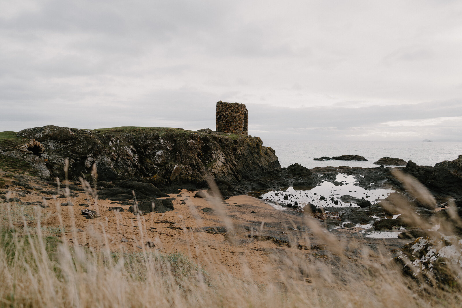 A ruined stone tower stands on a rocky headland, white sea foam breaking over rocks below and dune grass framing the foreground.