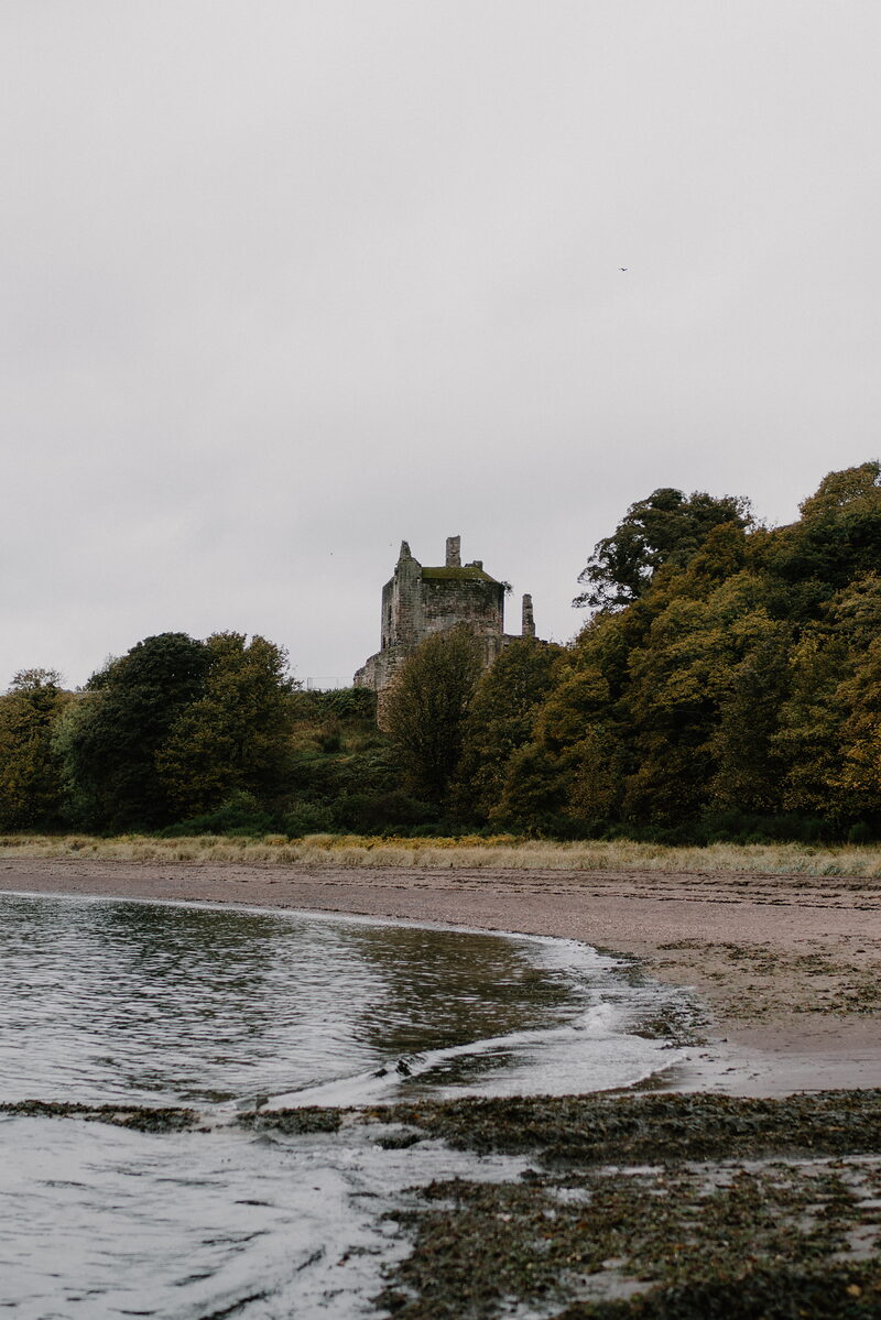 A ruined castle tower rises above autumn trees on a wooded shore, calm water and a sandy beach curving in front.