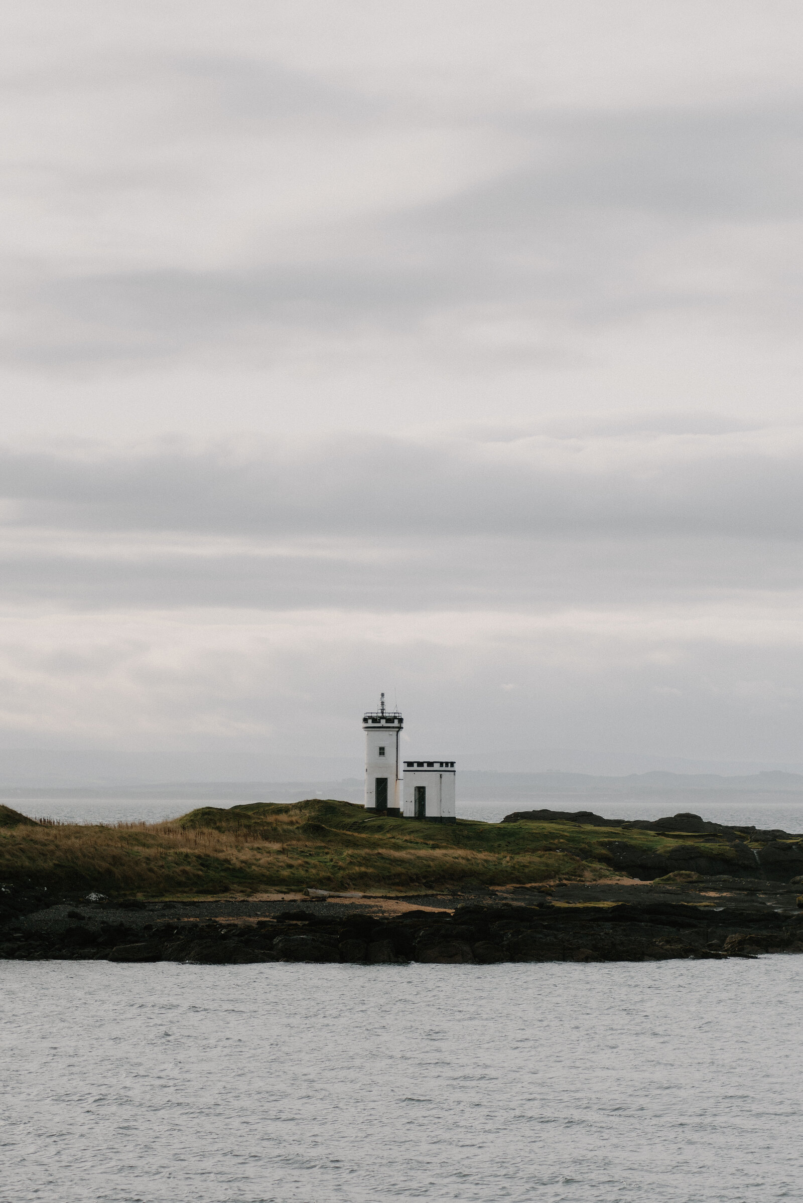 A white lighthouse with a black lantern room stands on a grassy rocky point, calm sea stretching around it.