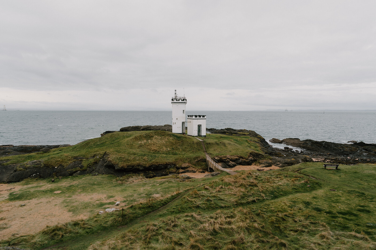 A white lighthouse with a black lantern room stands on a grassy point, a wooden bench nearby and calm sea stretching beyond.