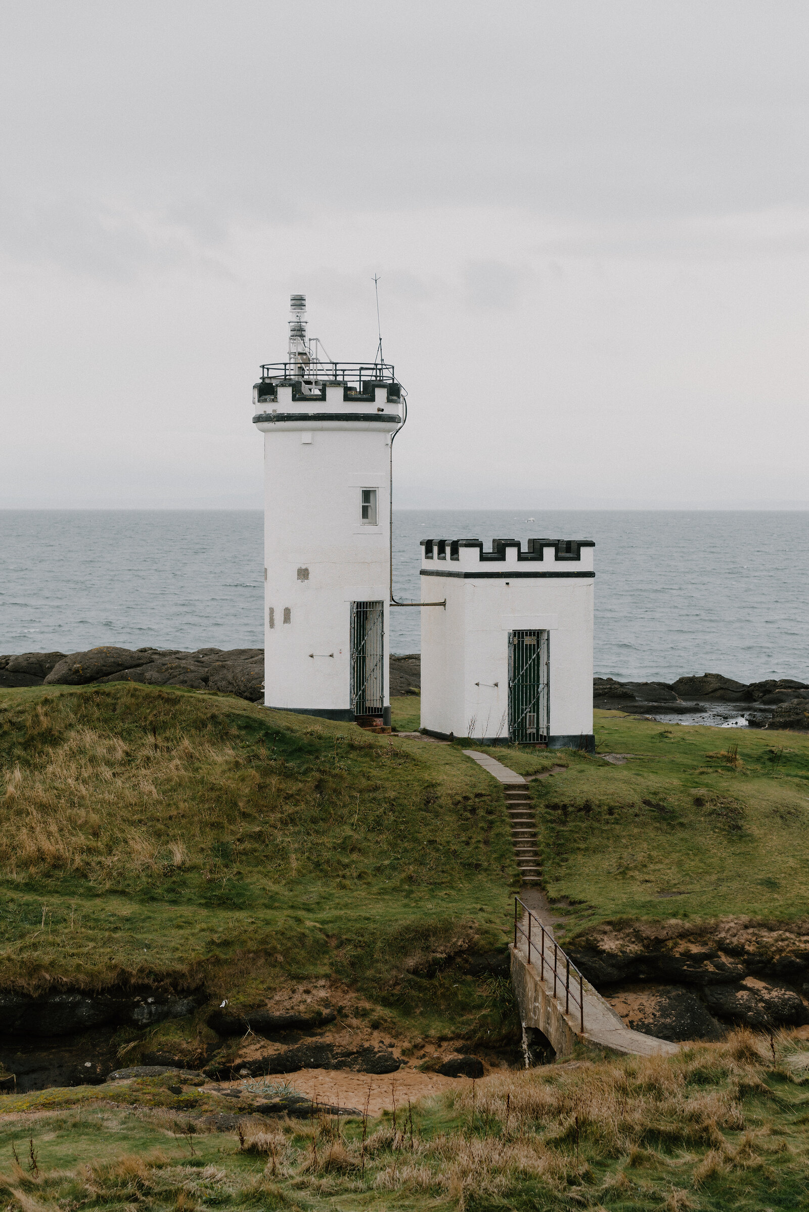 A white lighthouse stands beside a smaller crenellated tower on a grassy point, a footbridge leading across a gully towards them.