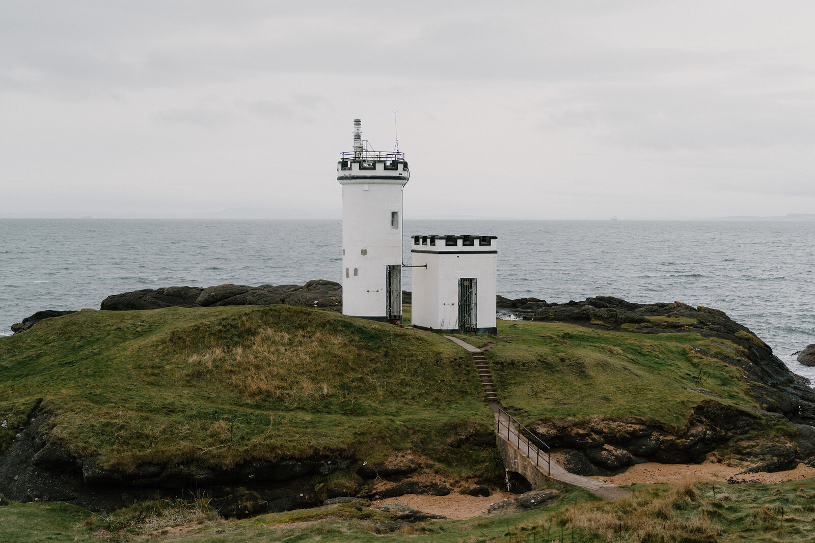 A white lighthouse stands on a grassy point above the sea, a stepped path and footbridge leading across a gully towards it.