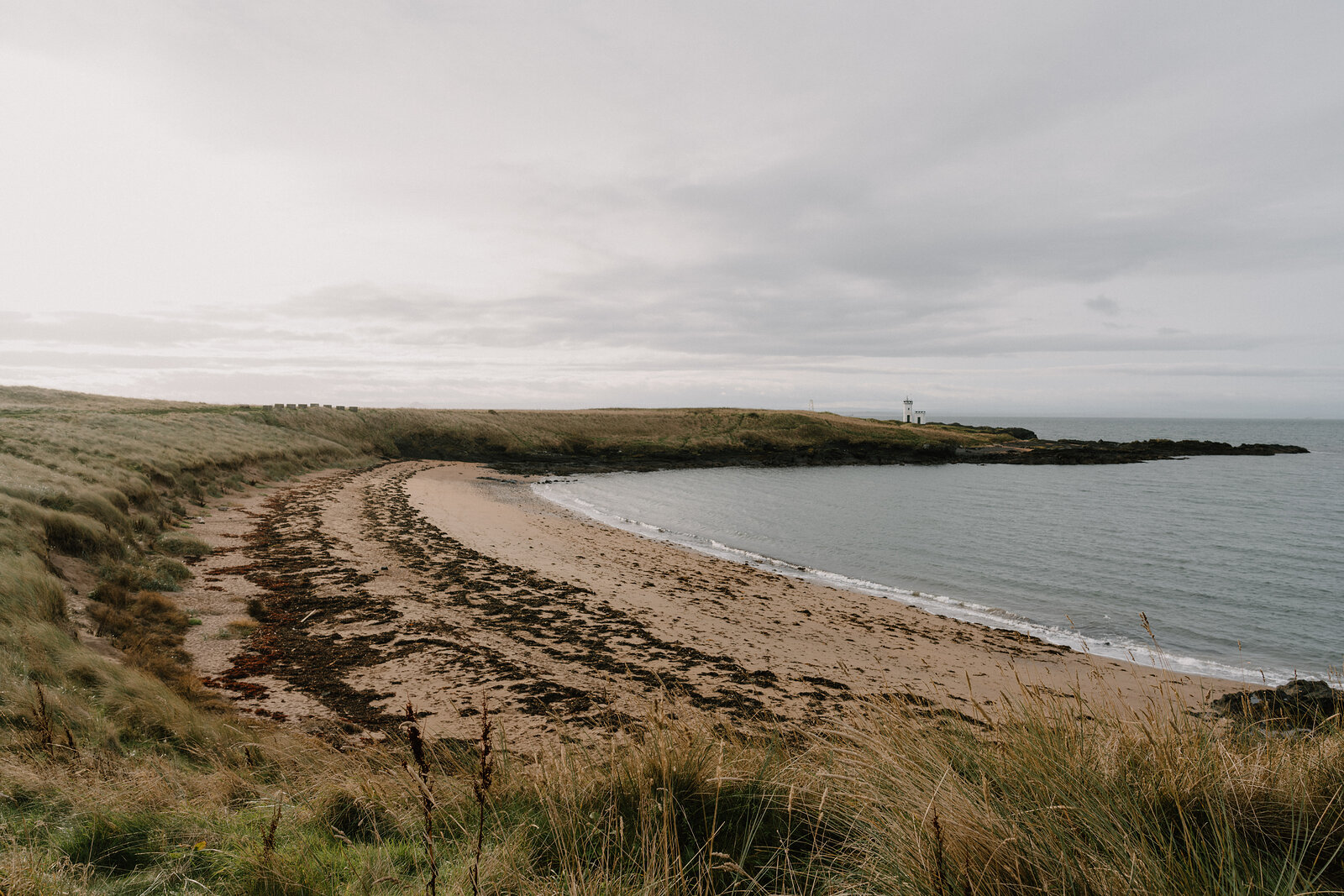 A sandy beach curves beneath dune grass, a distant lighthouse standing on a green headland at the far end.