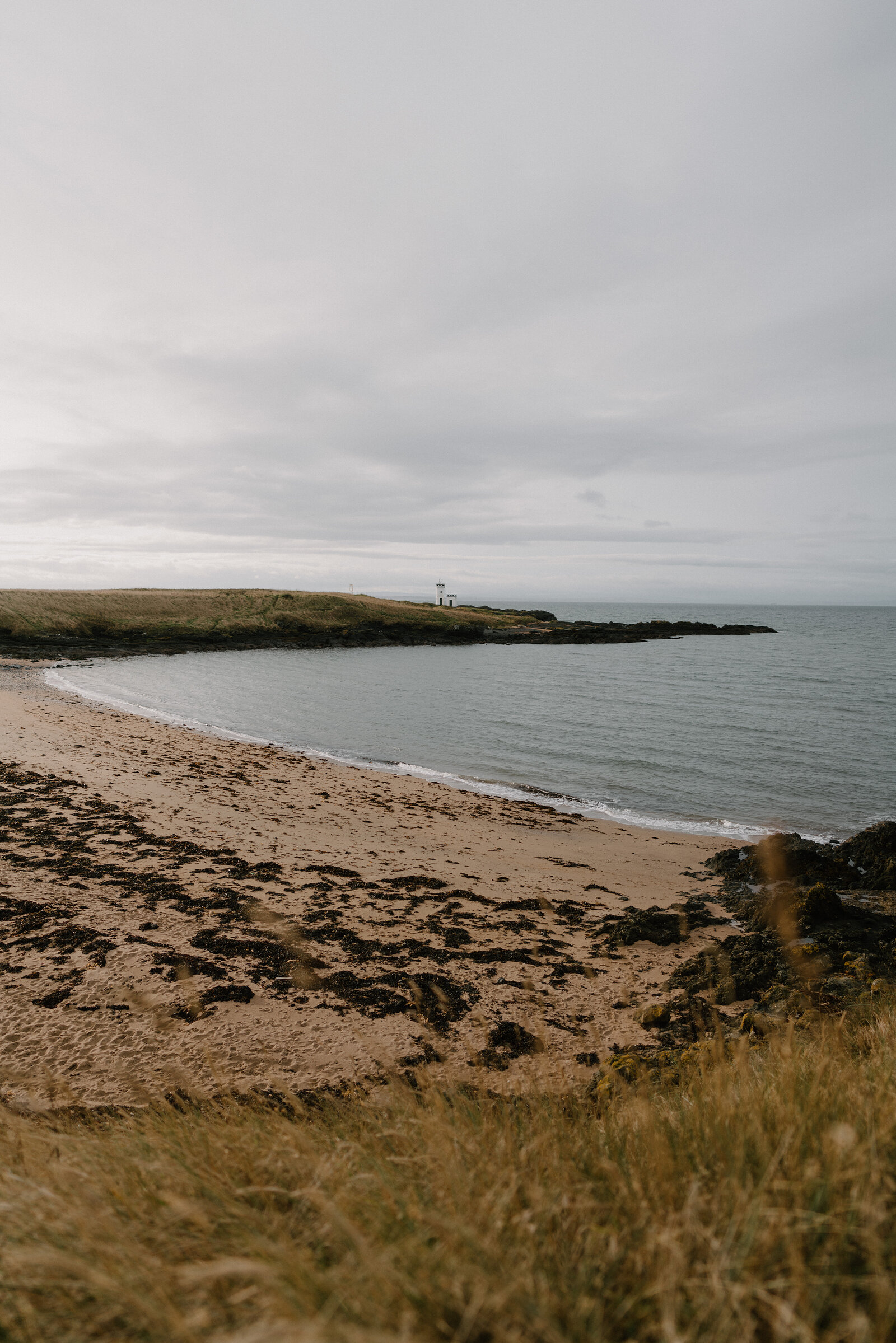 A sandy beach curves beneath dune grass, a distant lighthouse standing on a green headland at the far end.