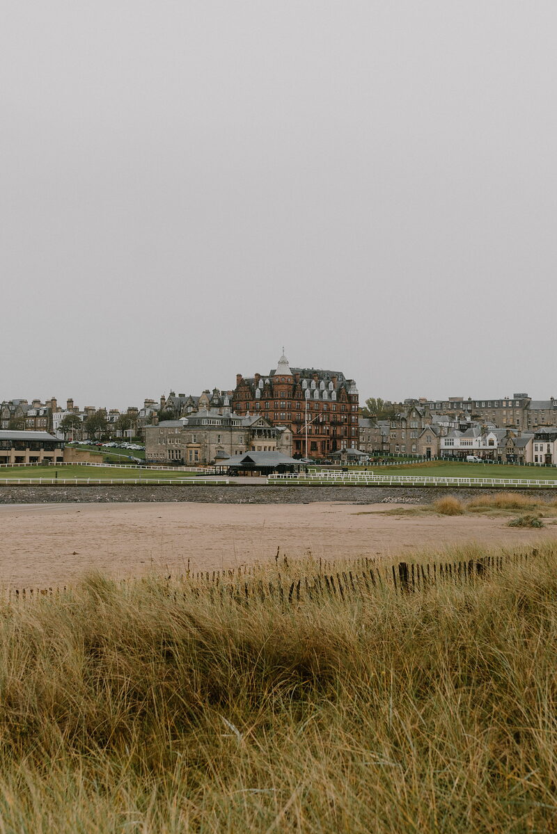 A red sandstone hotel with a domed corner tower rises above the rooftops of a coastal town, dune grass and a sandy beach in the foreground.