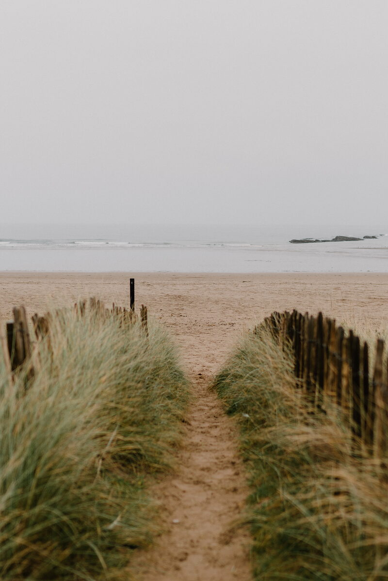 A sandy path flanked by wooden picket fencing and marram grass leads towards a beach with rocks offshore.