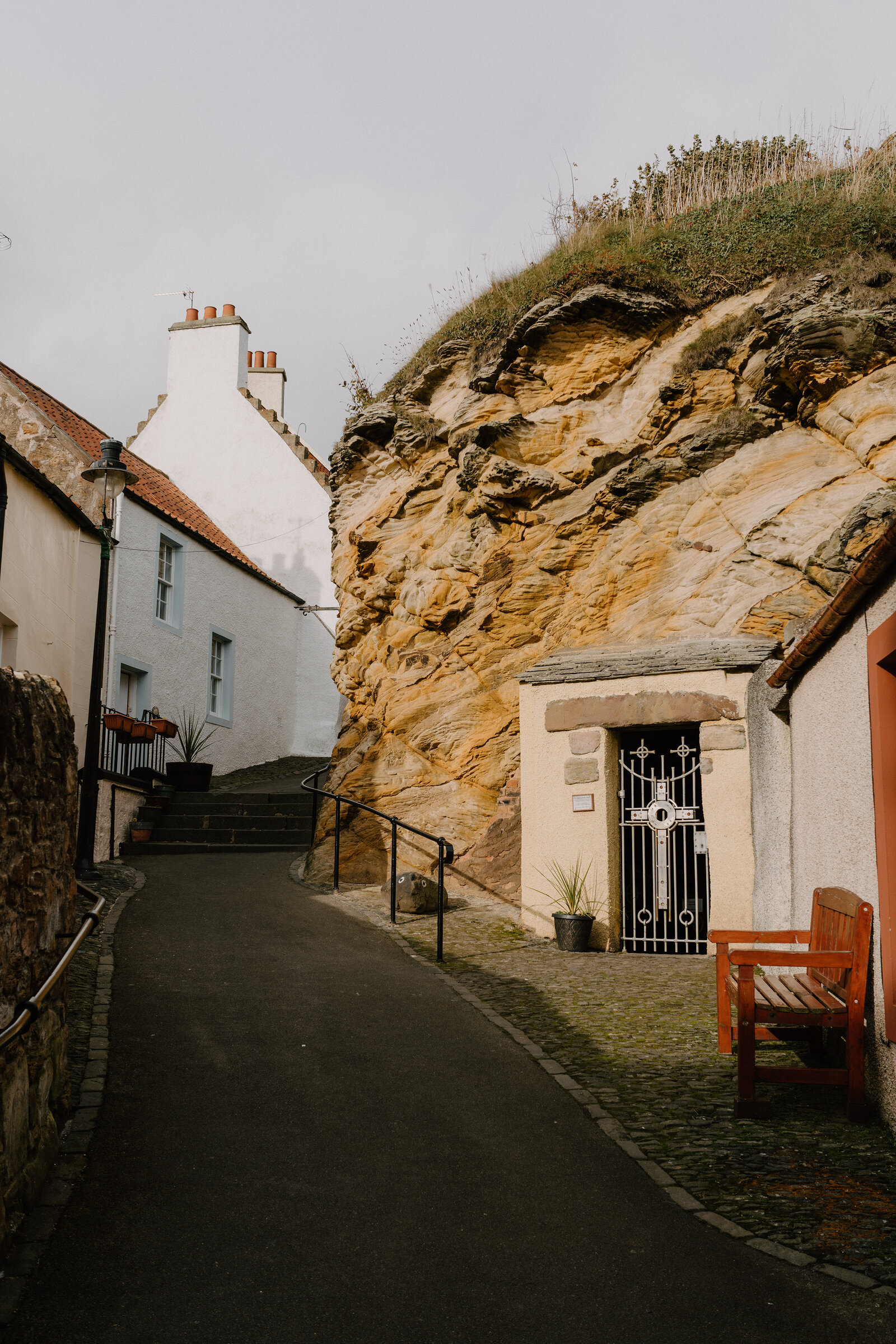 A narrow lane runs past whitewashed cottages towards a sandstone cliff face, a wrought iron gate set into an archway and a wooden bench standing nearby.