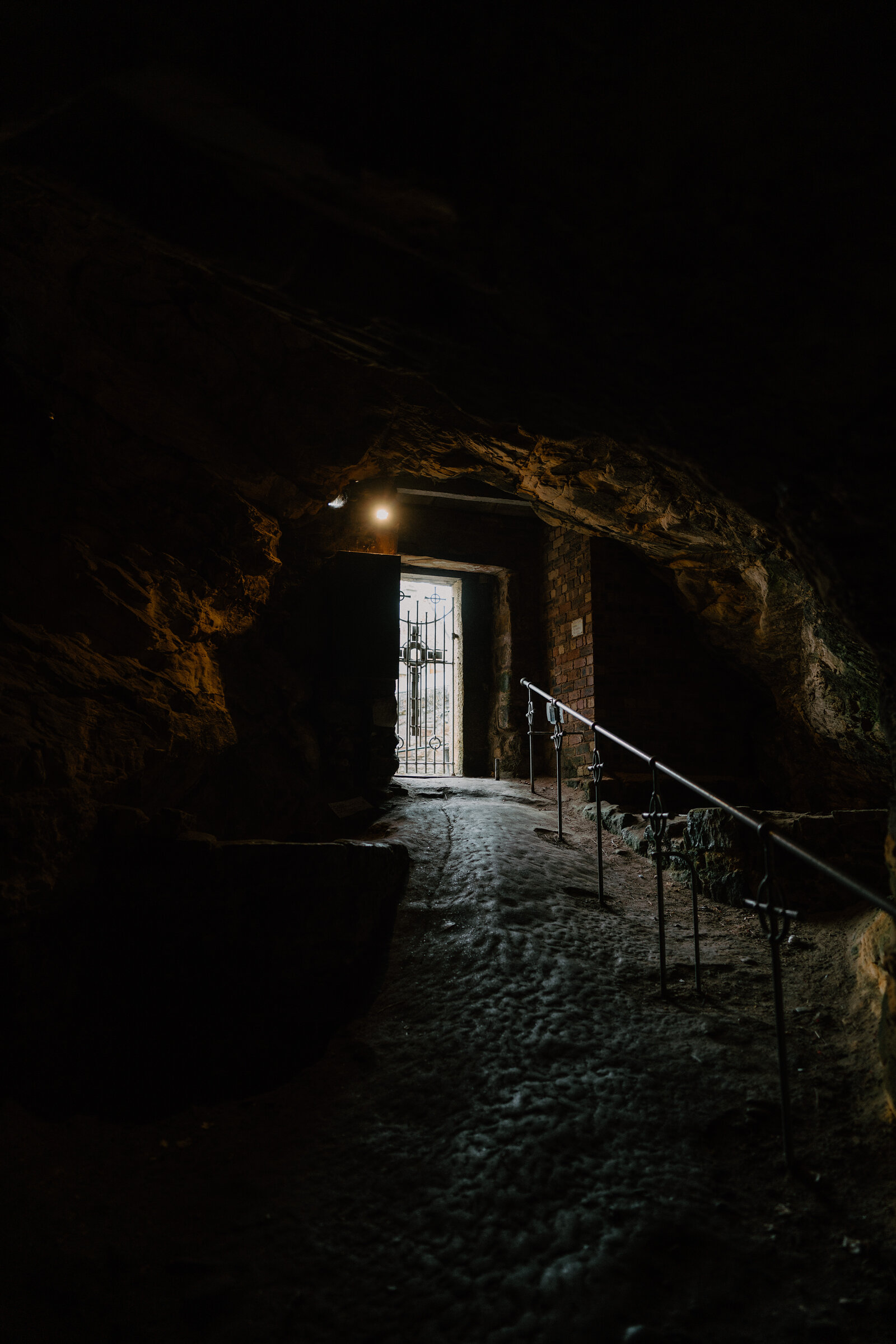 A rough-hewn rock passage leads towards a lit wrought iron gate, a handrail running along the uneven stone floor.