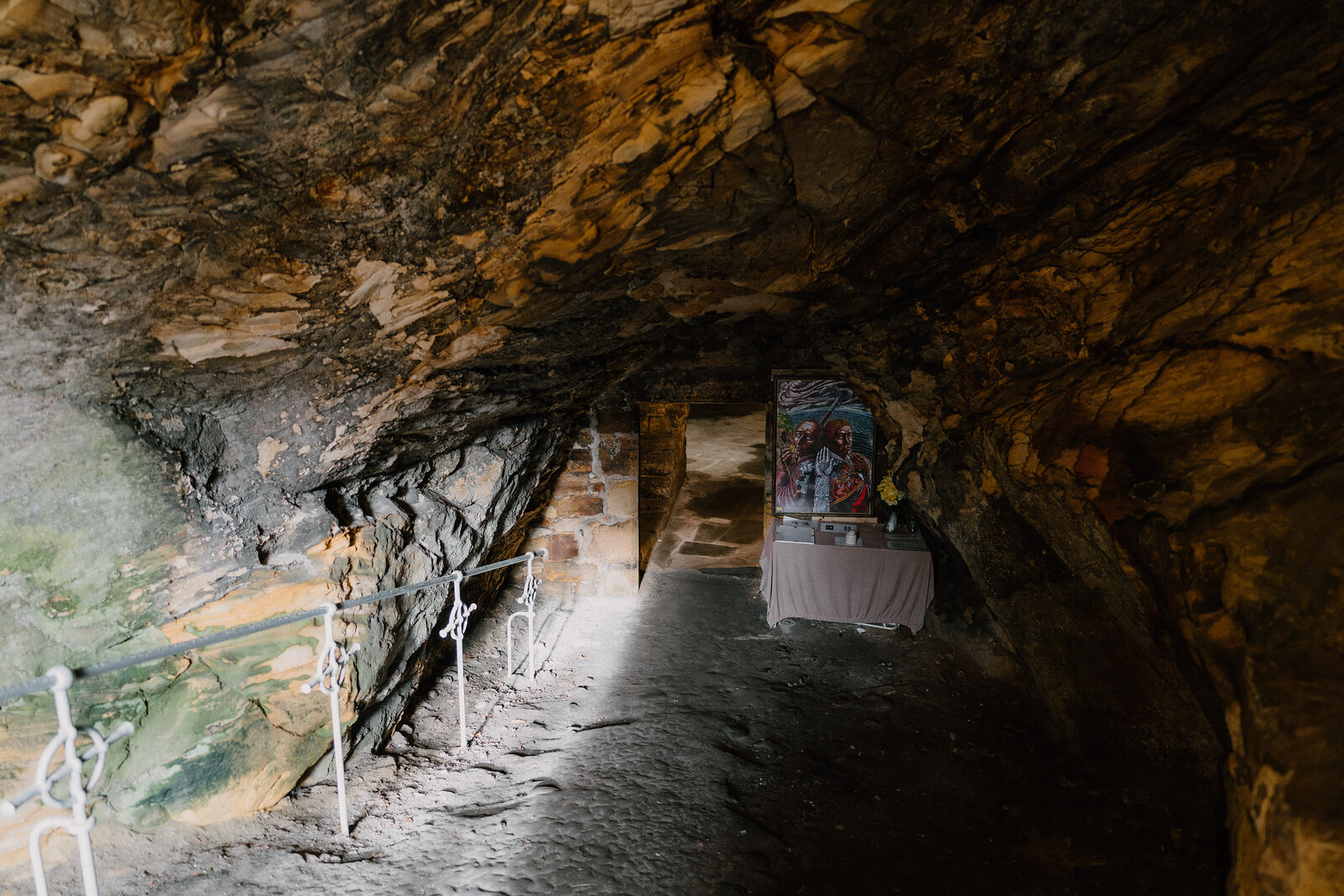 A rough rock-cut passage leads to a small lit shrine with a framed image, a metal handrail running along the uneven floor.