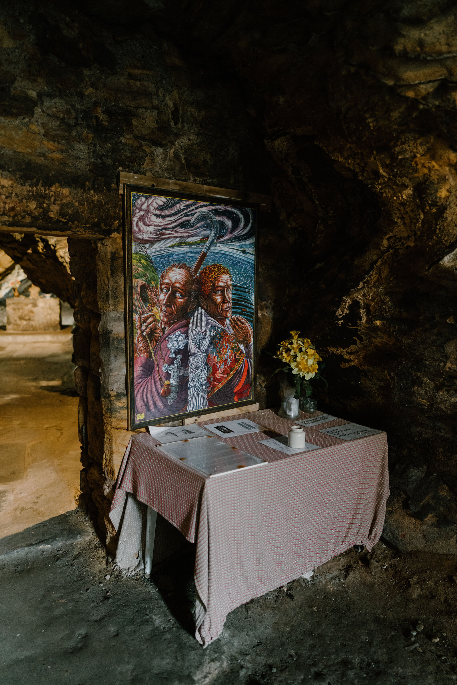 A framed painting of two robed figures hangs above a cloth-covered table set with a small vase of yellow flowers in a rock-cut shrine.