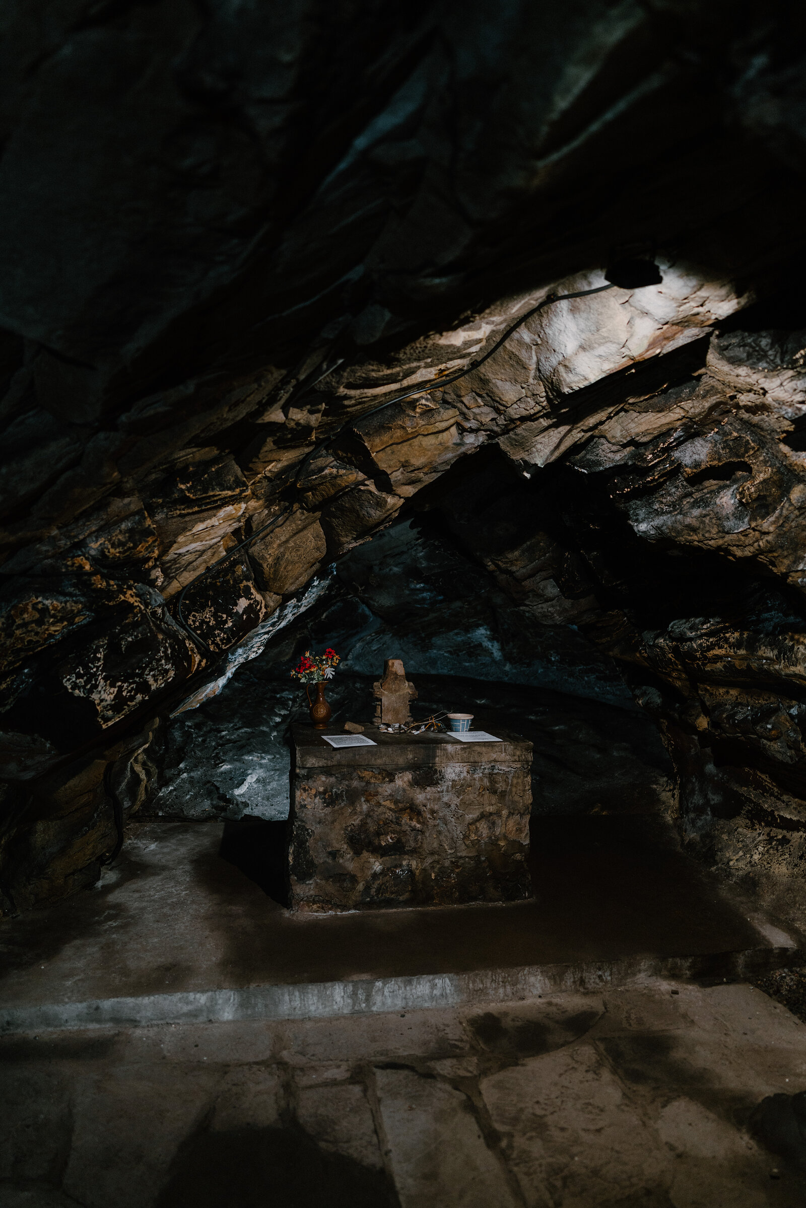 A small stone altar holds a carved figure, flowers and a card in a dark rock-cut chamber, faint light falling from an opening above.