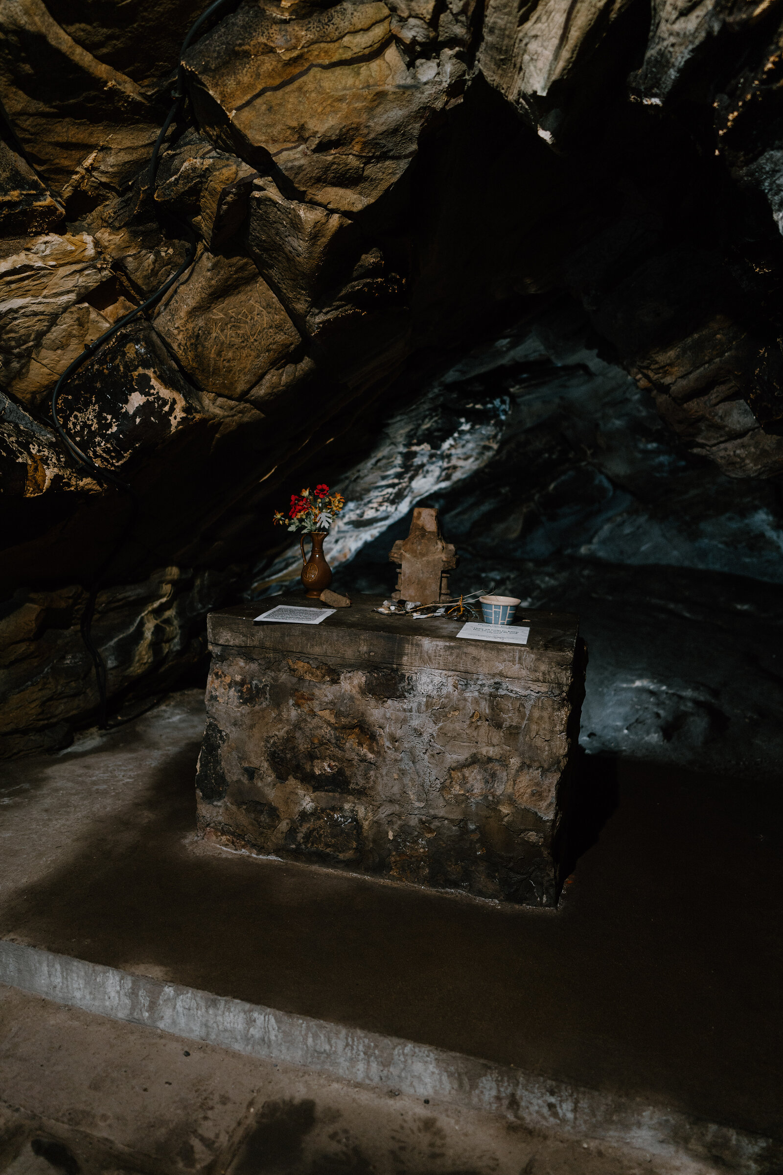 A small stone altar holds a carved figure and a vase of flowers in a dark rock-cut cave, faint daylight falling from an opening above.