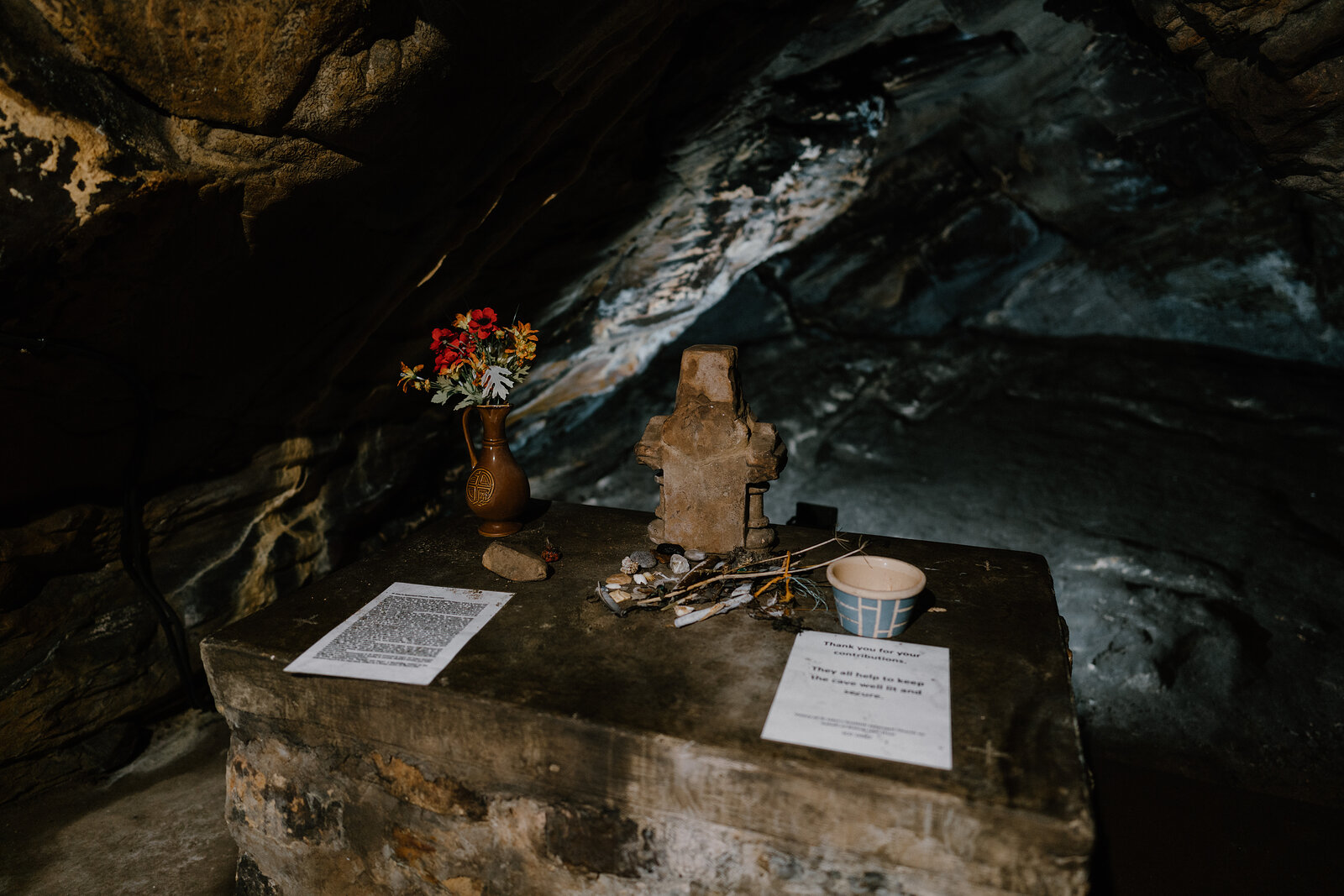 A stone altar in a rock-cut cave holds a small cross, a jug of flowers and a candle, handwritten notices propped on either side.