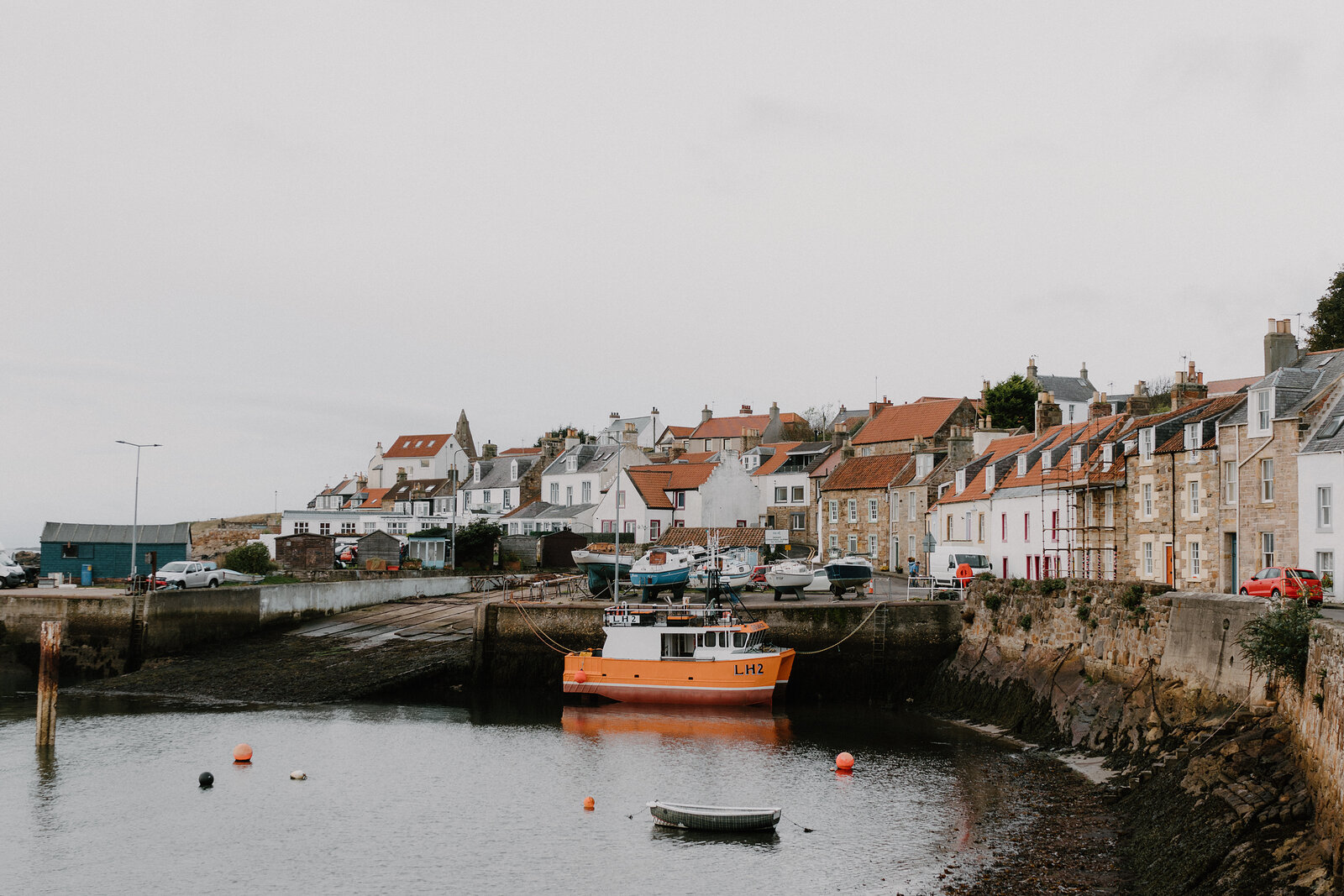 An orange fishing boat rests in a stone harbour at low tide, a village of white and red-roofed houses rising behind the seawall.