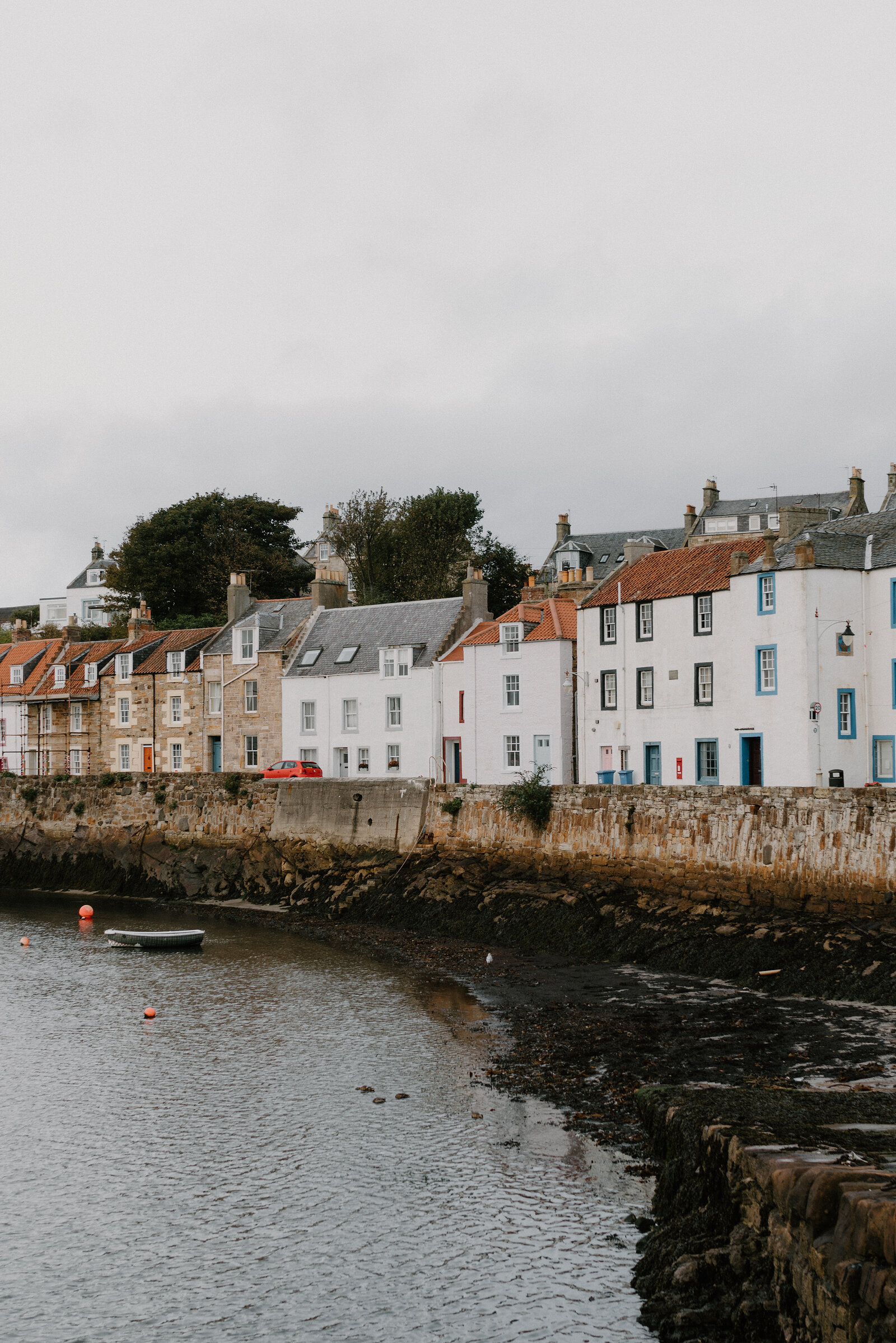 A row of white, cream and blue-trimmed houses lines a stone harbour wall, a small boat resting on the mud at low tide.