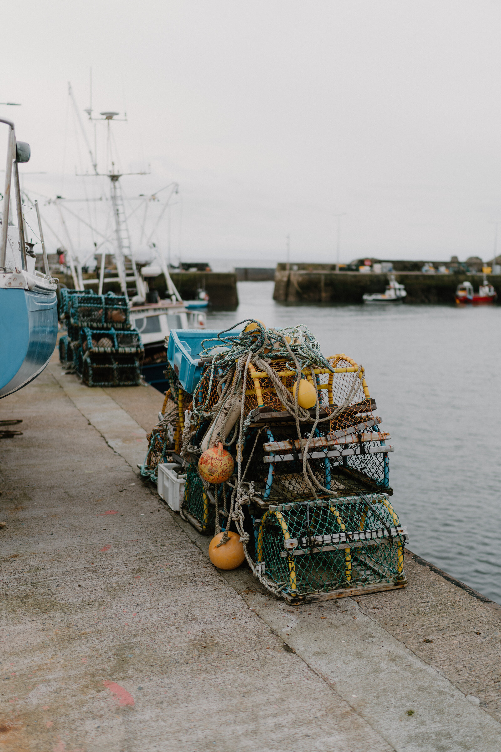 Stacked lobster creels and coiled rope sit on a harbour quay beside moored boats, a breakwater visible across the water.