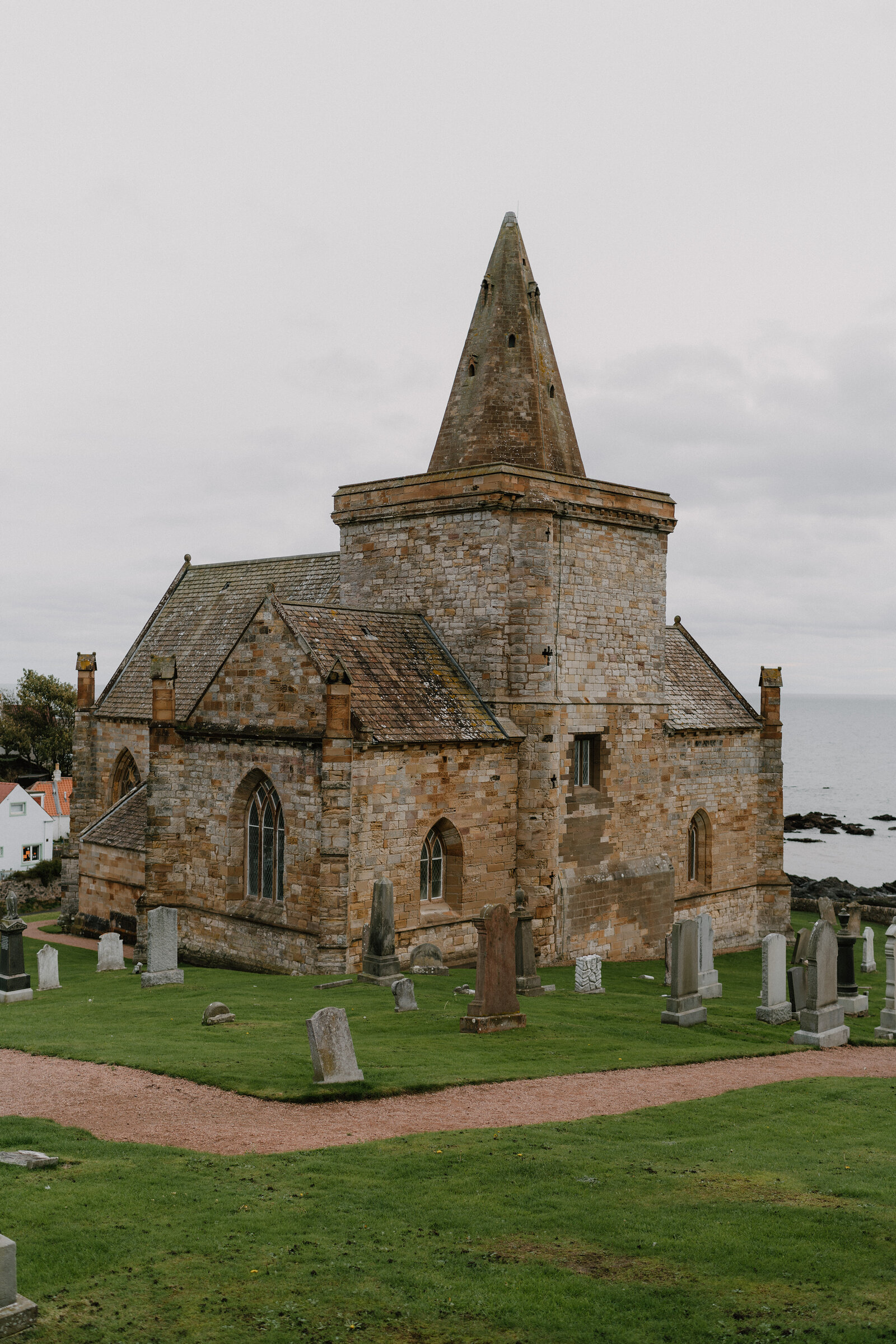 A stone church with a conical spire stands above a graveyard of weathered headstones, the sea visible just beyond the wall.