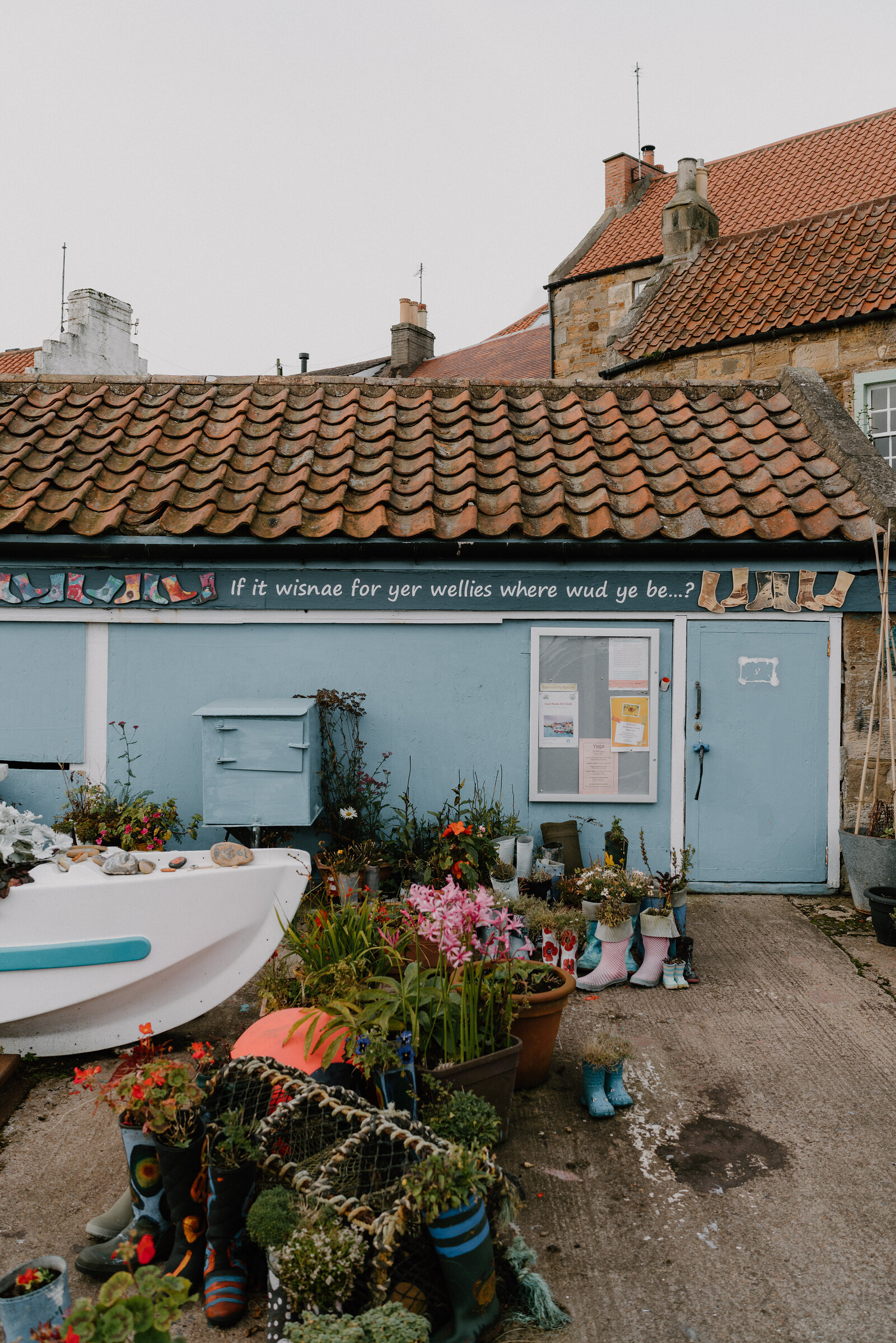 A pale blue shed with a pantiled roof is painted with a mural reading "If it wisnae for yer wellies where wud ye be...?", planted wellington boots and a small boat filled with flowers standing outside.