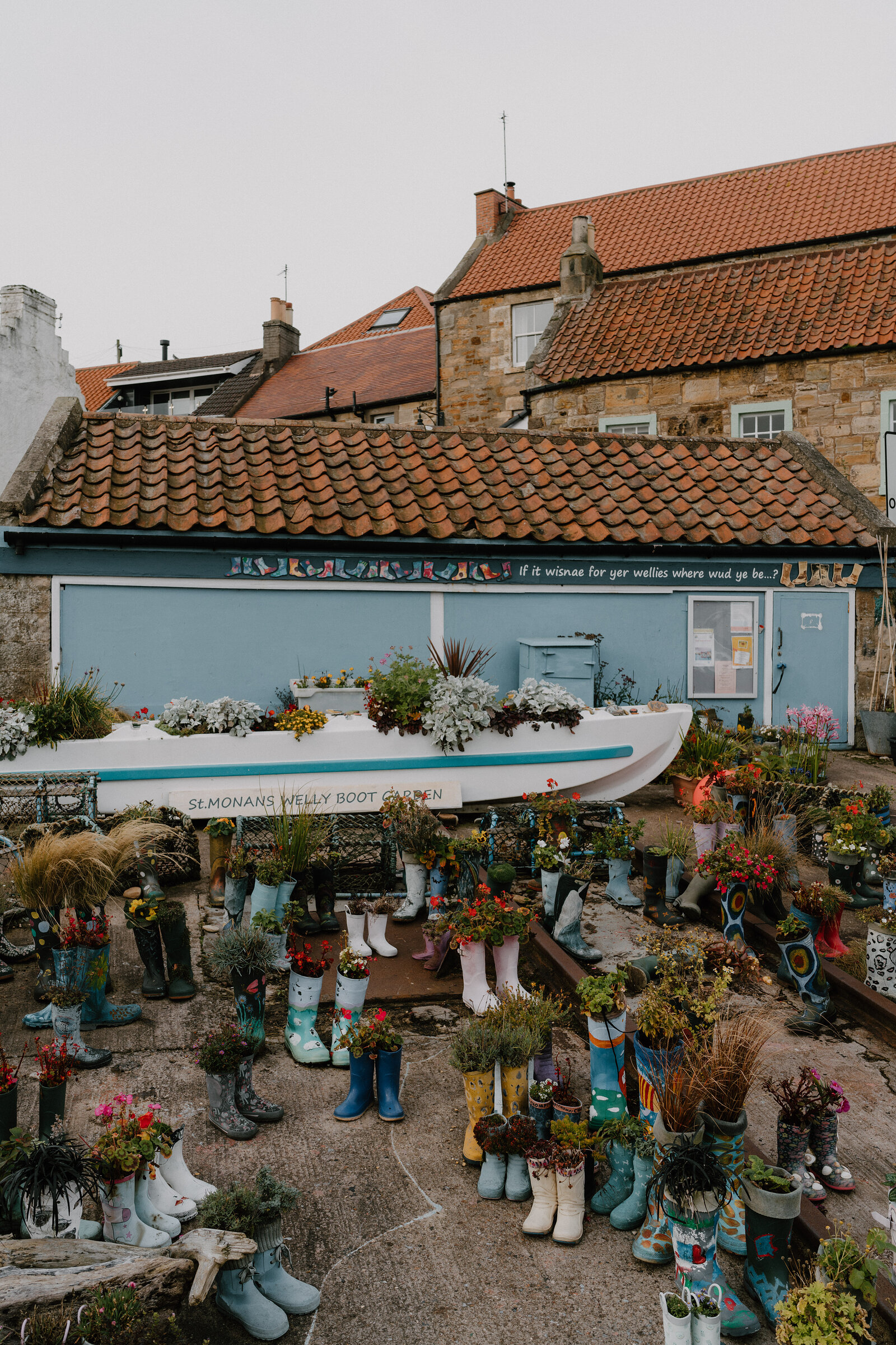 Rows of wellington boots planted with flowers fill a courtyard beneath a mural reading "if it wisnae for yer wellies where wud ye be...?", a boat planted with flowers standing among them.