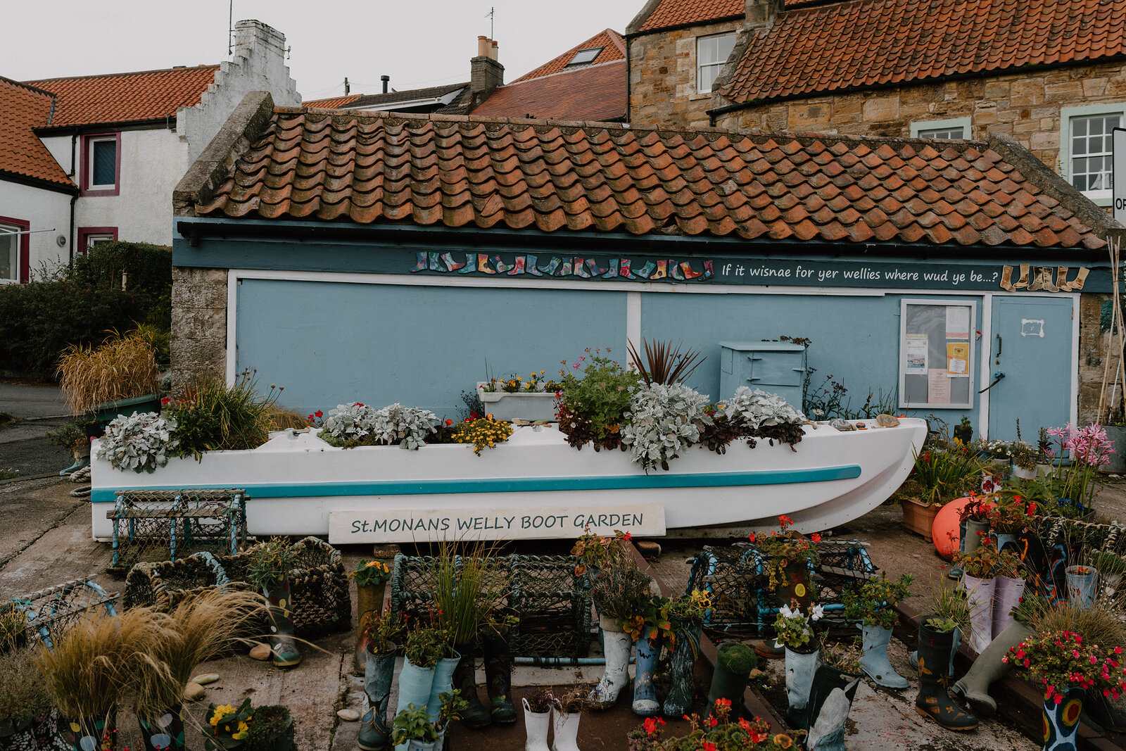 A small boat filled with flowers stands before a pale blue shed painted with a mural reading "if it wisnae for yer wellies where wud ye be", wellington boots planted with flowers lining the ground.