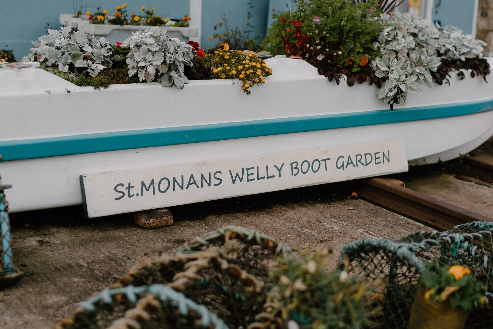 A wooden name plate reading "St.Monans Welly Boot Garden" is fixed to the side of a boat planted with flowers, lobster creels arranged on the ground nearby.