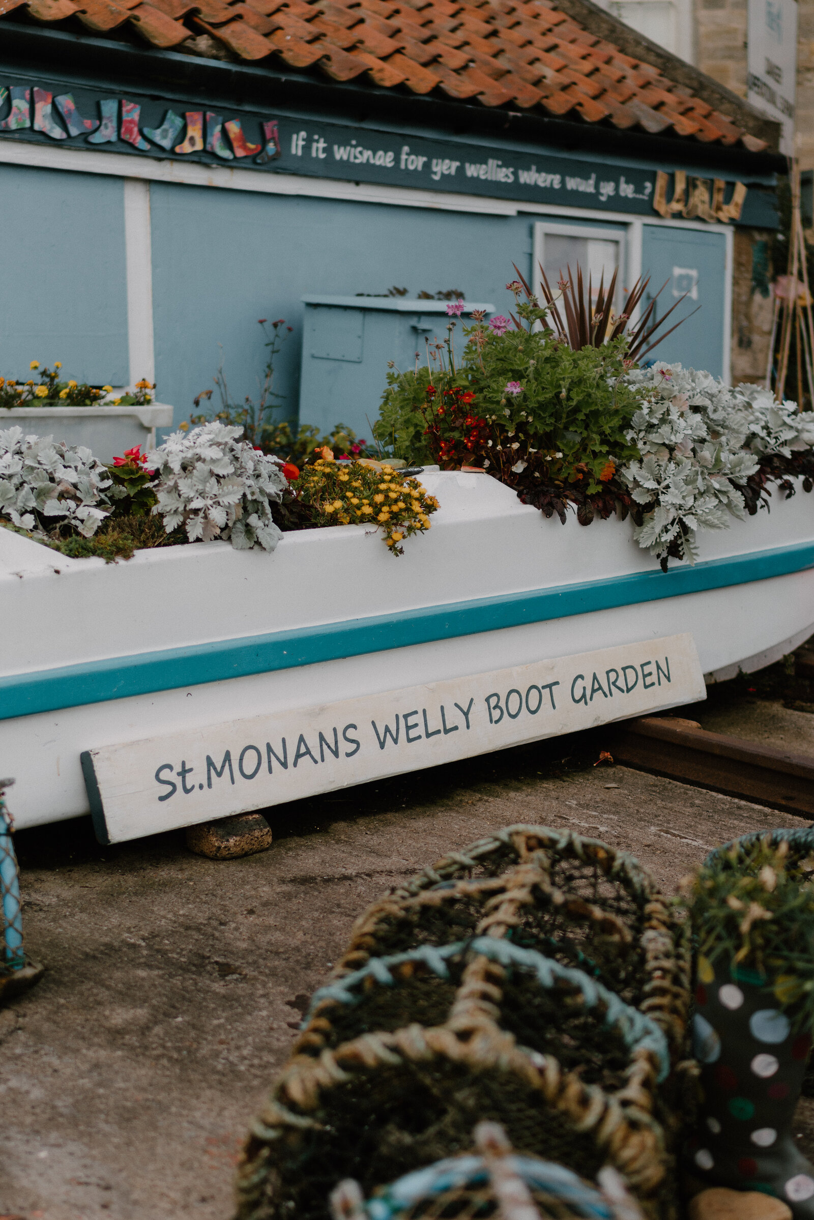 A boat planted with colourful flowers bears a sign reading "St.Monans Welly Boot Garden", lobster creels and patterned wellington boots arranged in front of it.