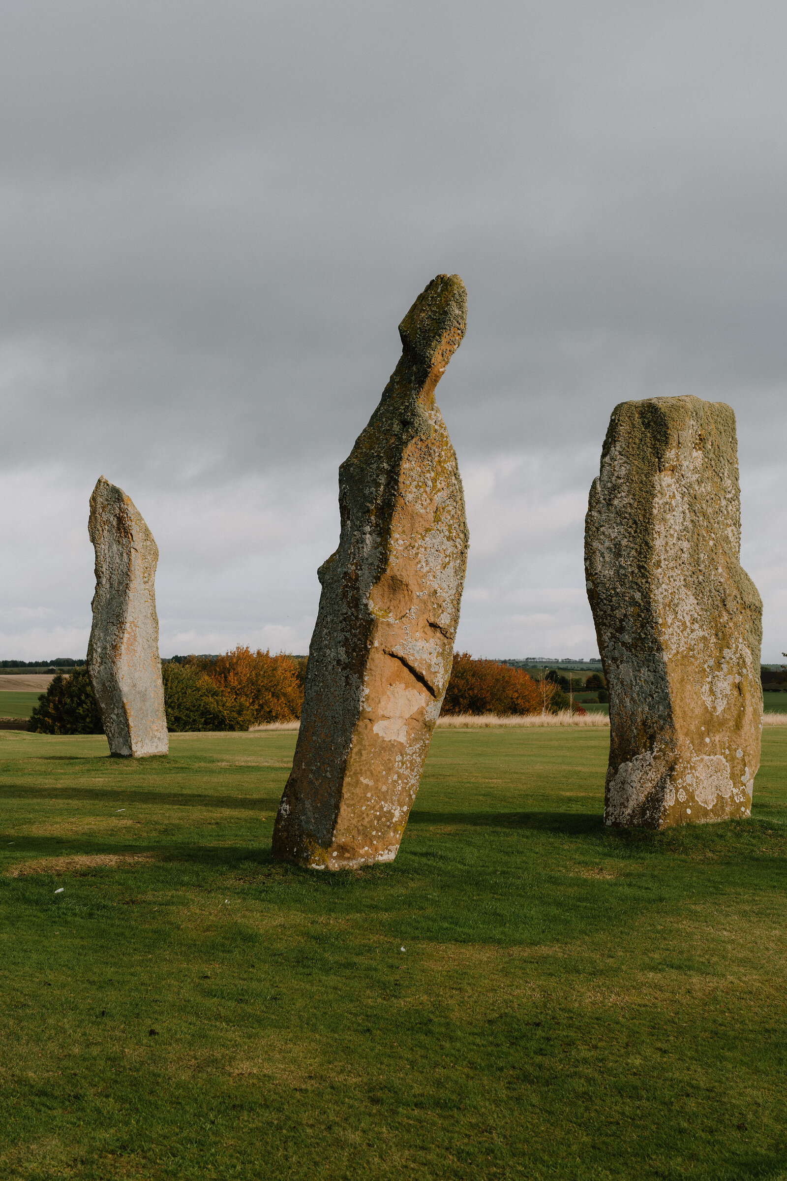 Three tall standing stones rise from a mown field beneath a cloudy sky, hedgerows and a distant field visible in the background.