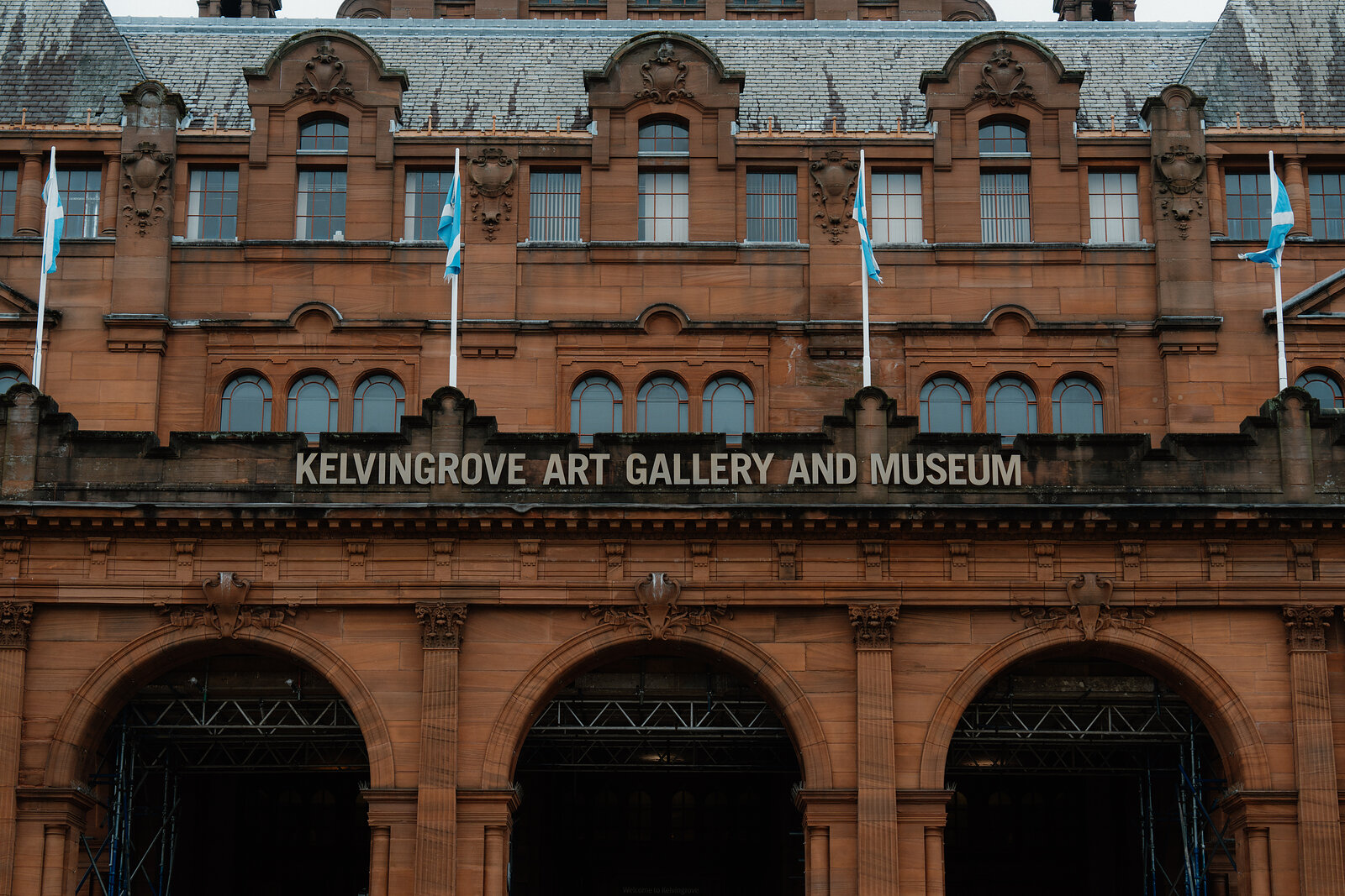 A red sandstone gallery building carries a sign reading 'Kelvingrove Art Gallery and Museum' between rows of arched windows and decorative flags.