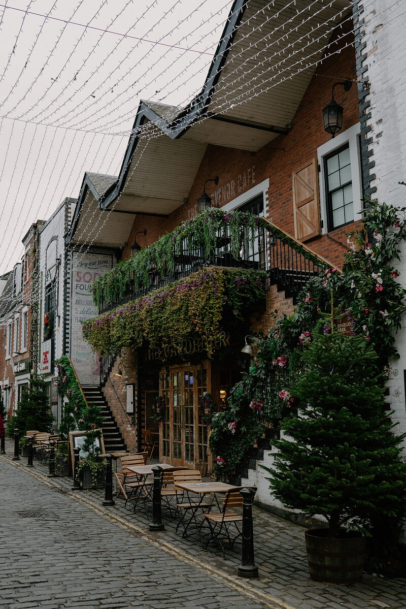 A brick building with a balcony draped in trailing plants and flowers stands along a cobbled lane strung with festoon lights, outdoor tables and small potted trees set below.