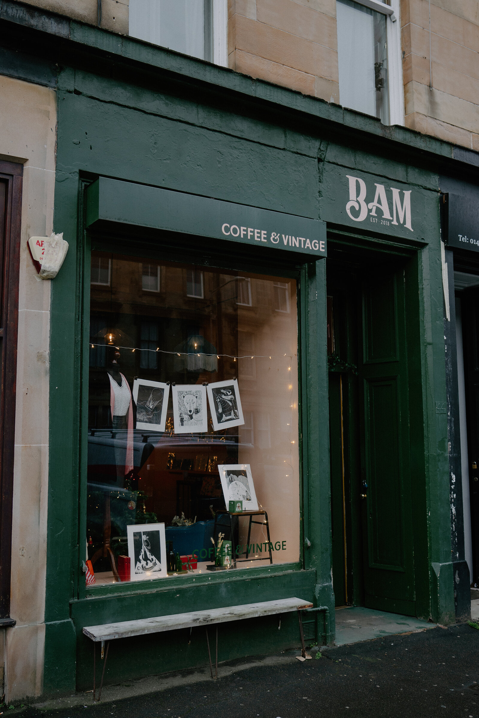 A dark green shopfront reads "BAM, coffee and vintage" beside a window displaying framed illustrations and vintage items, a bench standing outside.