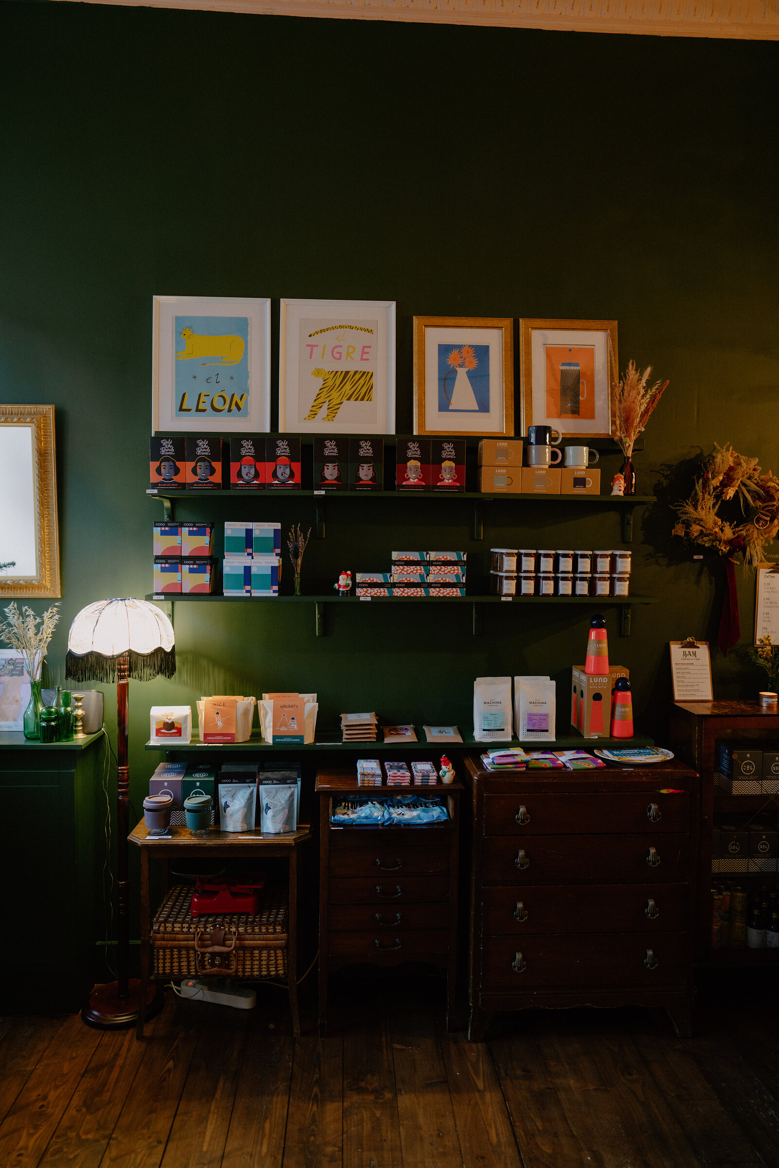 Framed illustrated prints hang above a chest of drawers and shelving of gift items against a dark green wall, a floor lamp standing to one side.