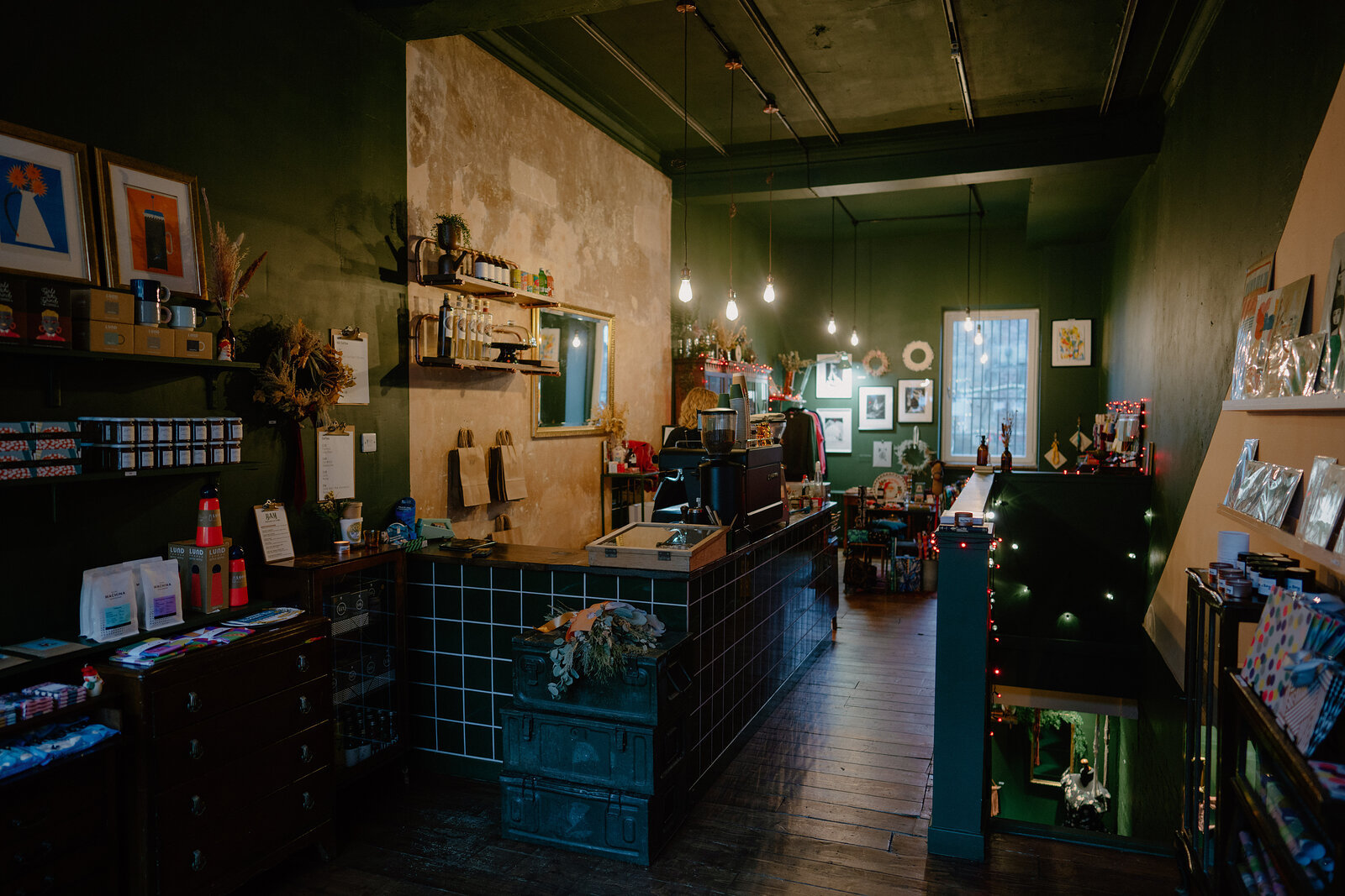A tiled shop counter stands beneath pendant lights and a wreath, shelving of gift items and framed art lining the dark green walls.