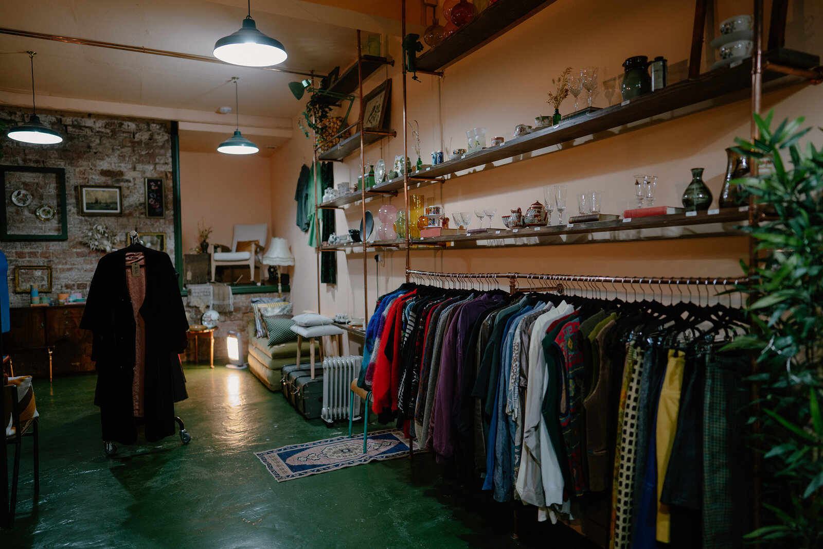 Racks of vintage clothing fill a shop with green flooring, shelving of glassware and framed pictures on the pink walls behind.