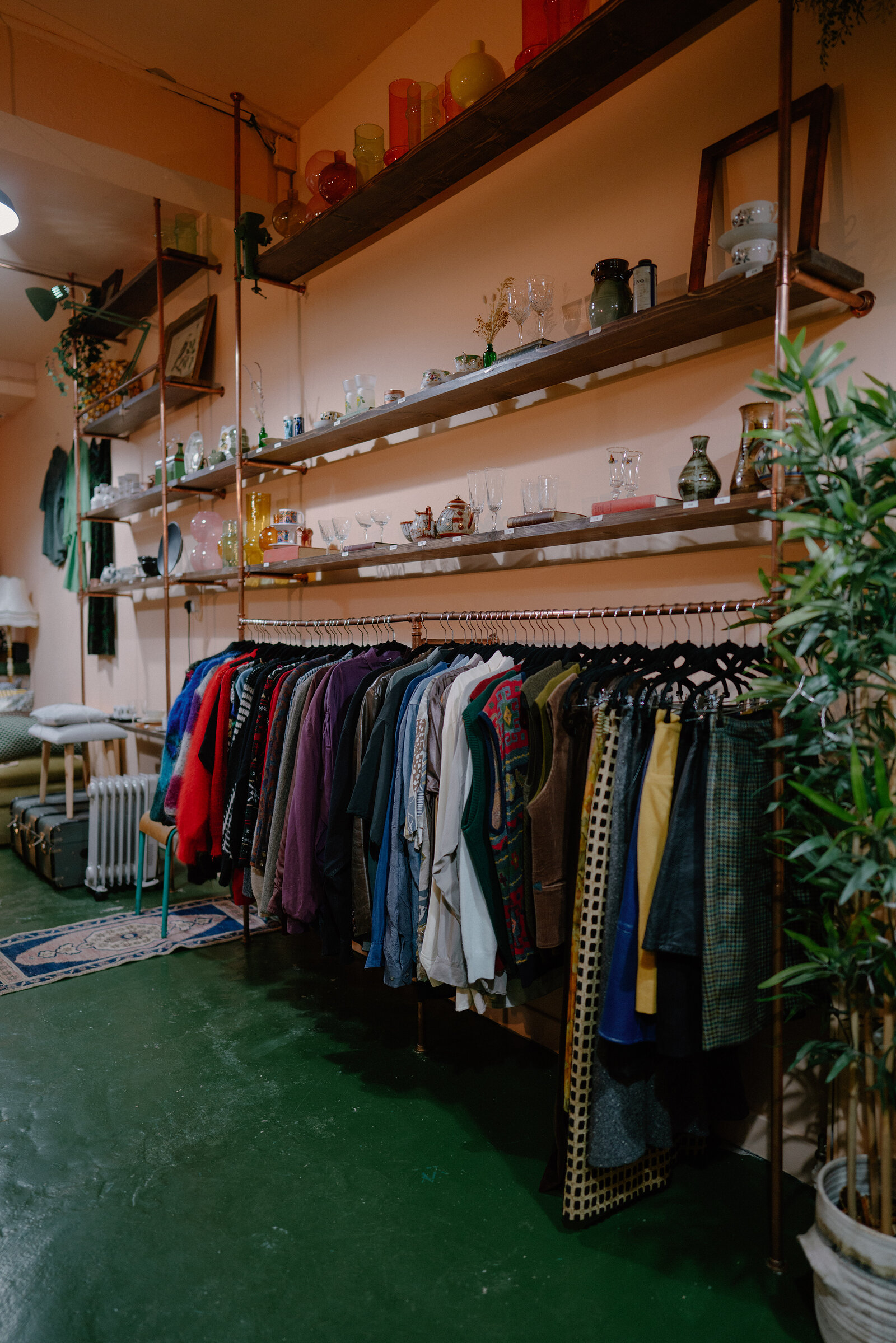 A rail of vintage clothing stands beneath shelving of glassware and ceramics against a pink wall, a green floor and potted plant furnishing the shop.
