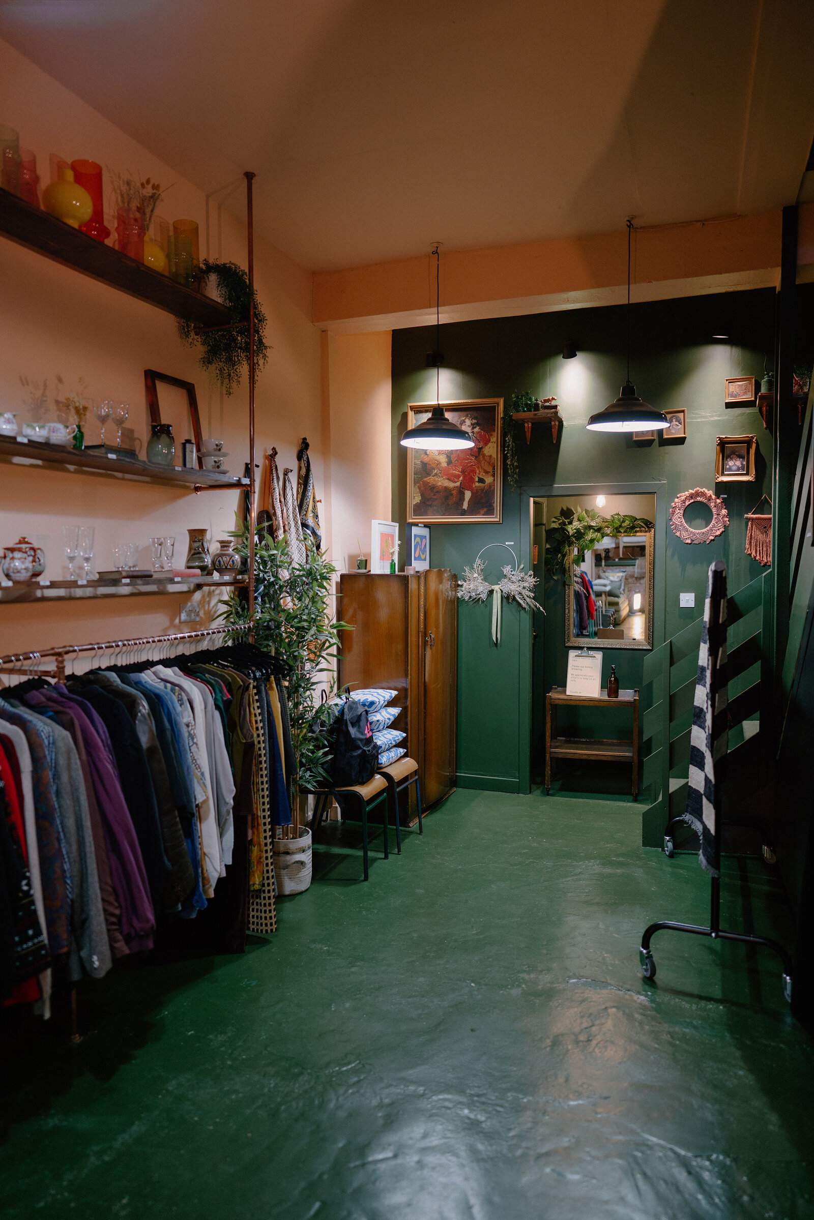A clothing rail and shelving of glassware fill a vintage shop with pink walls, a dark green corridor and framed pictures visible beyond.