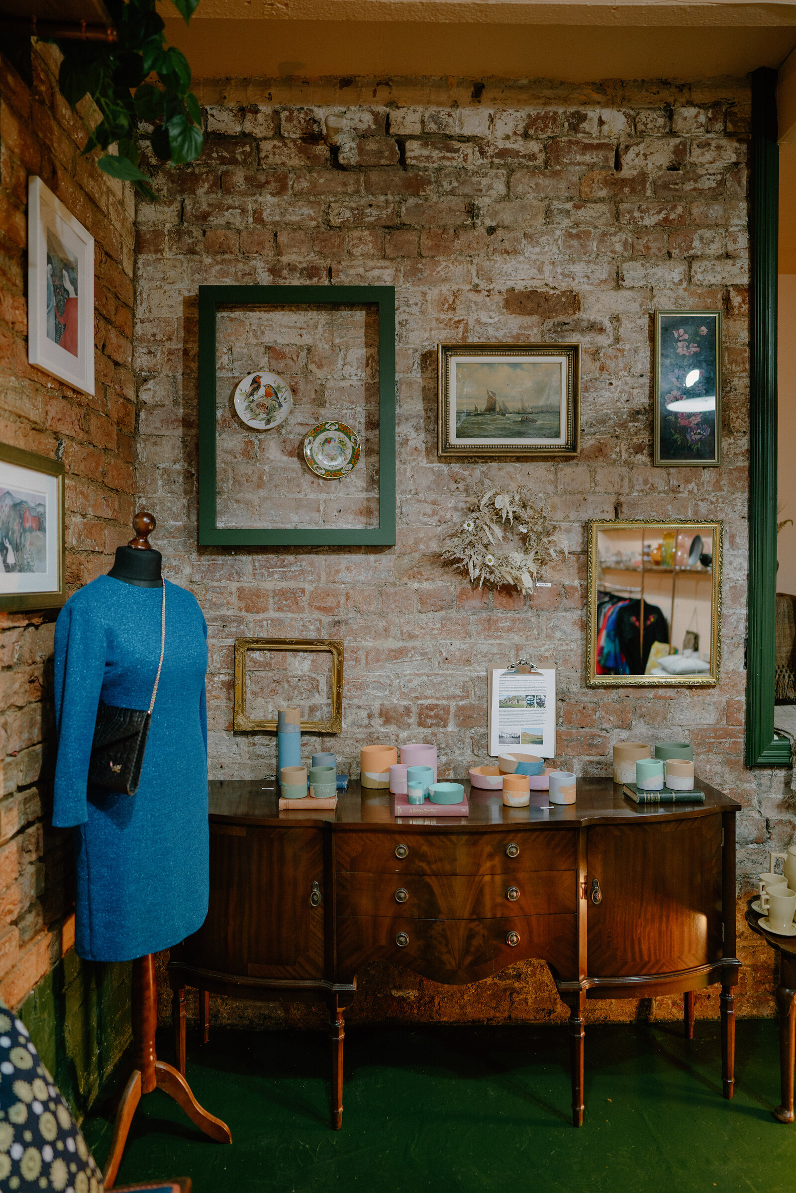 An exposed brick wall hung with mismatched framed pictures and empty frames rises above an antique sideboard set with pastel-coloured pots, a mannequin in a blue dress standing nearby.