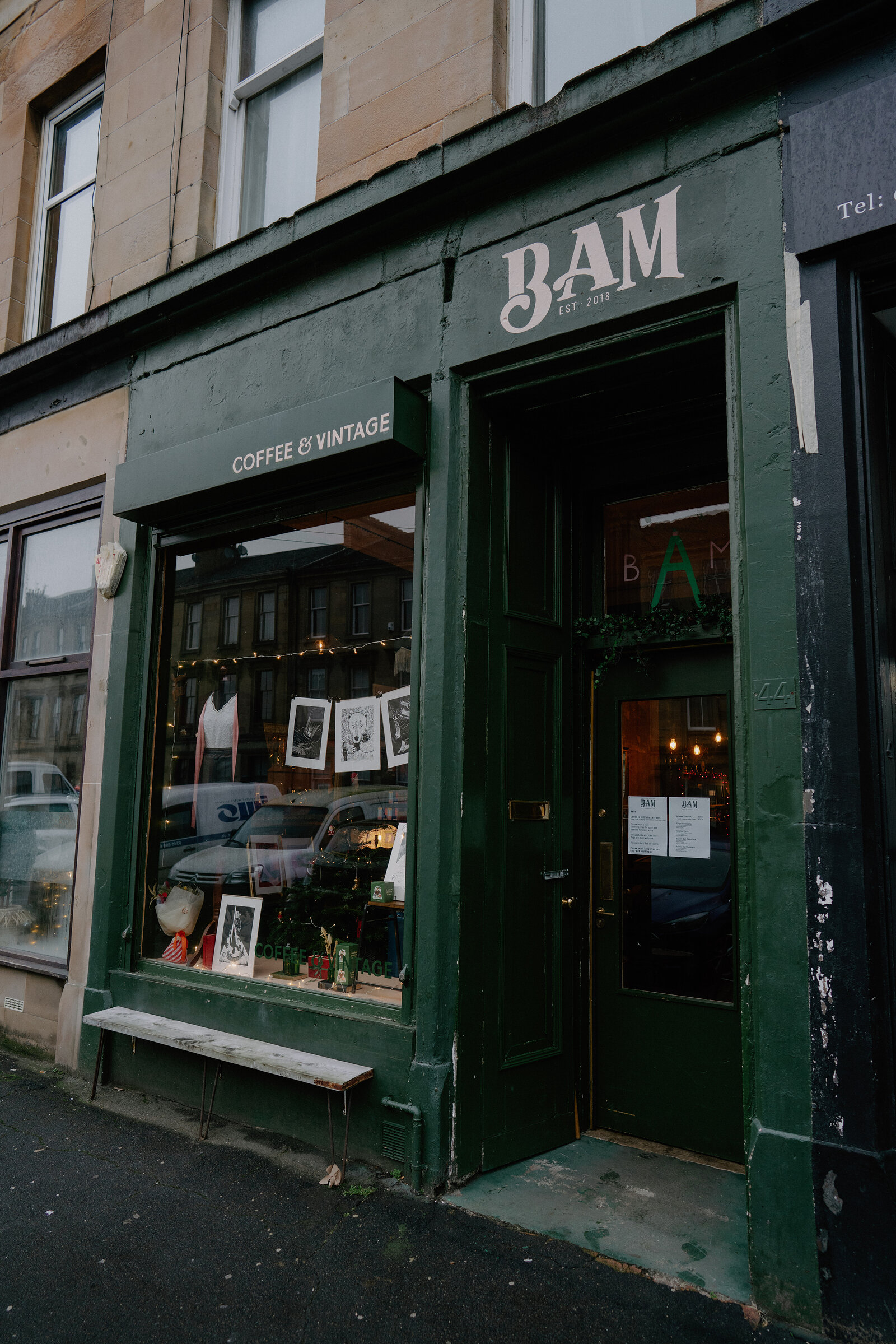 A dark green shopfront reads "BAM, coffee and vintage" beside a bench and window display of vintage items on a tenement street.
