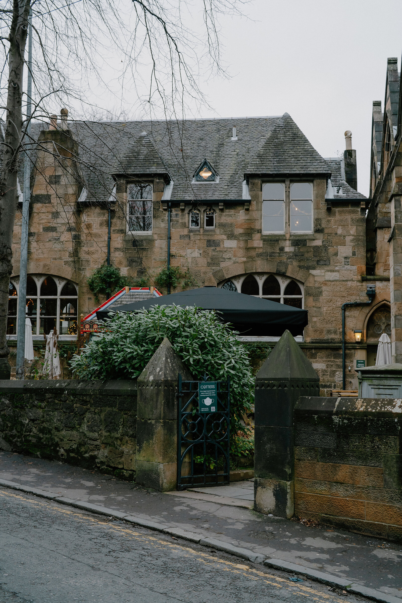 A Gothic sandstone building with arched windows stands behind a stone gateway marked "Cottiers", parasols and greenery filling the courtyard.