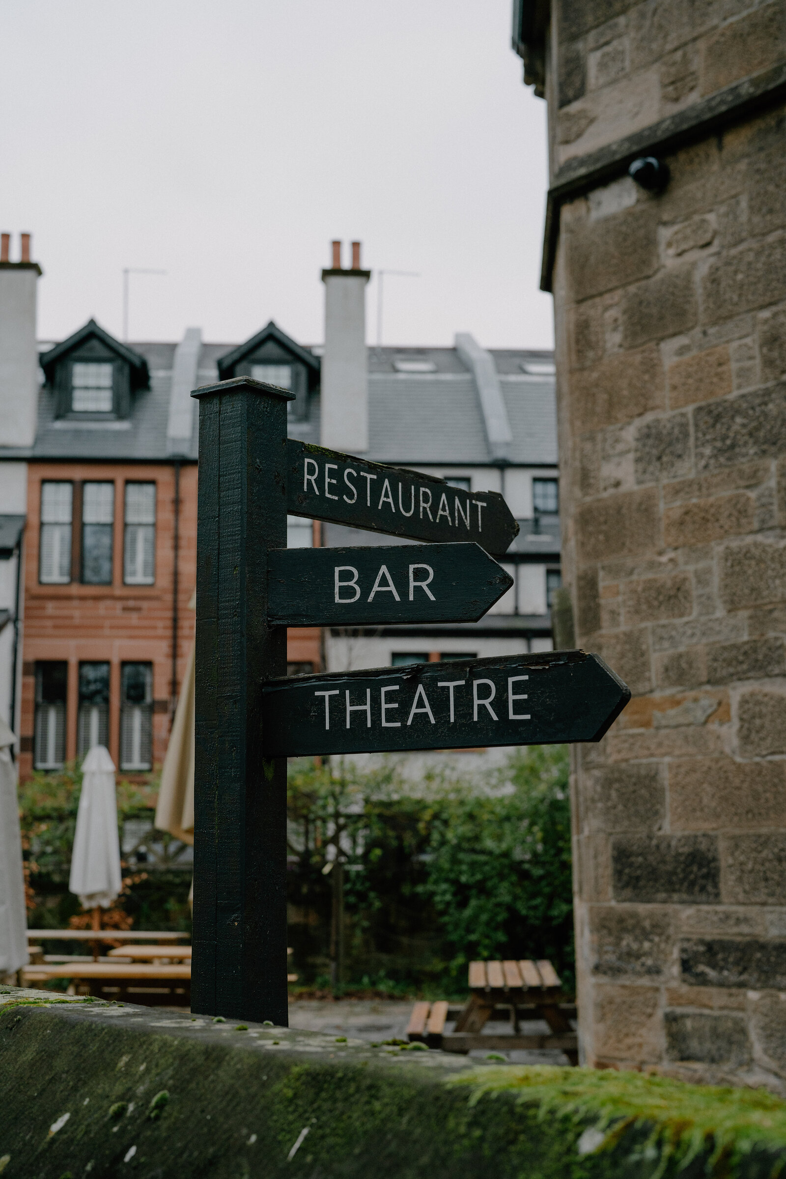 A black signpost with arrows reading "Restaurant", "Bar" and "Theatre" stands beside a sandstone wall, tenement buildings and picnic tables visible behind.