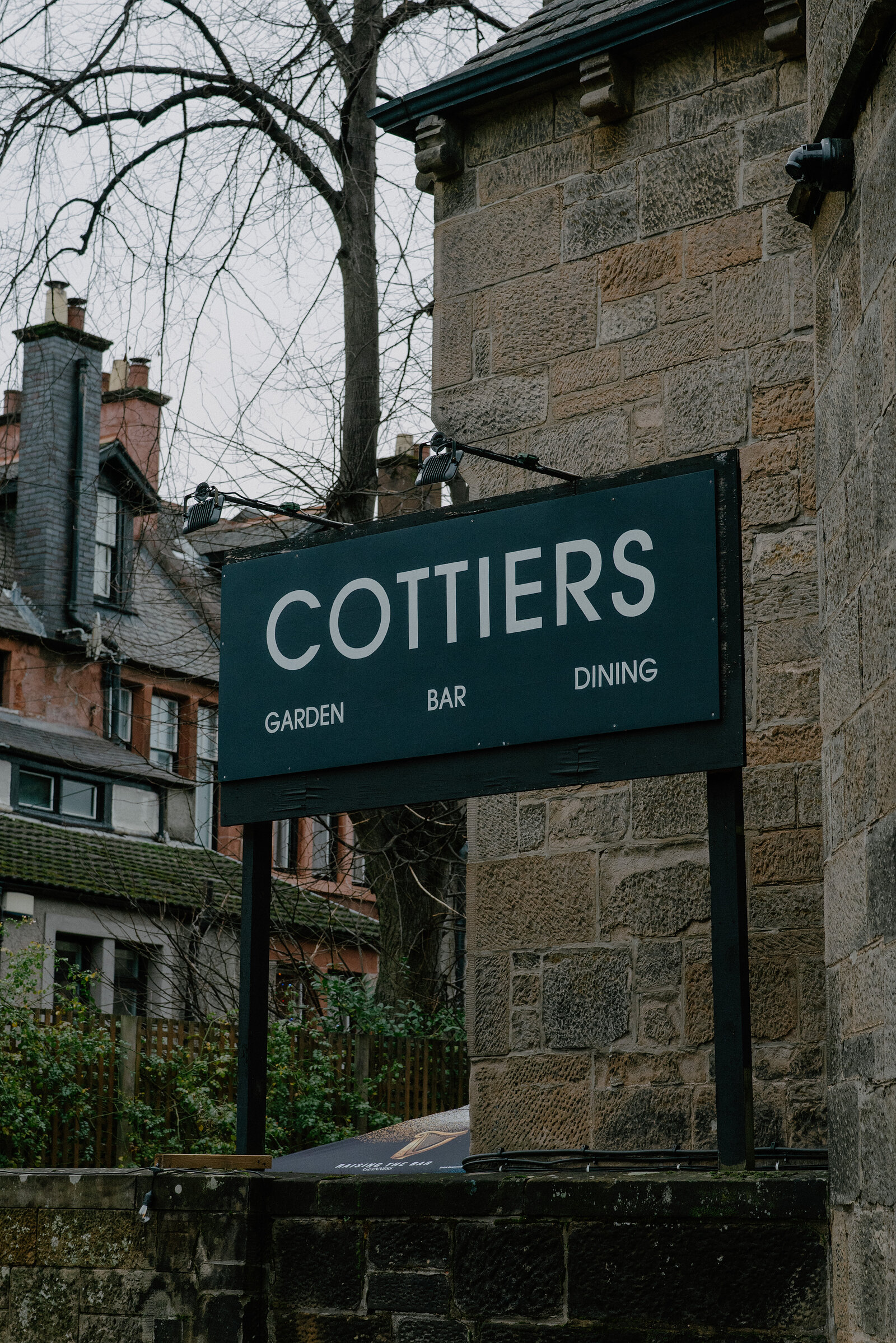 A sign reading "Cottiers, garden, bar, dining" is mounted on a stone wall, tenement buildings and bare trees behind.