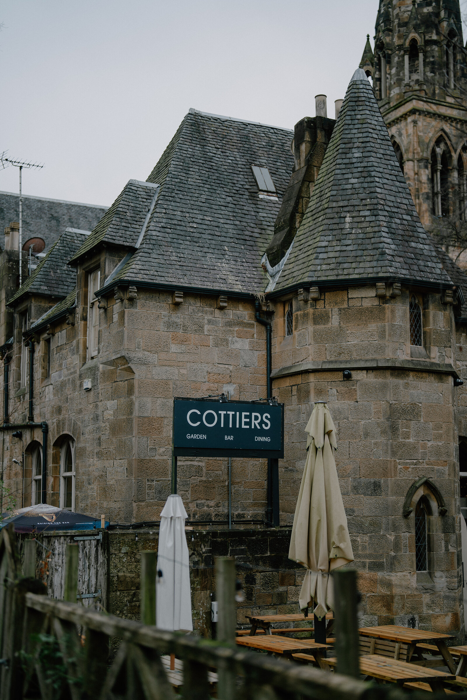 A sign reading "Cottiers, garden, bar, dining" is mounted on a turreted stone building, folded parasols and picnic tables standing in the courtyard below a church spire.